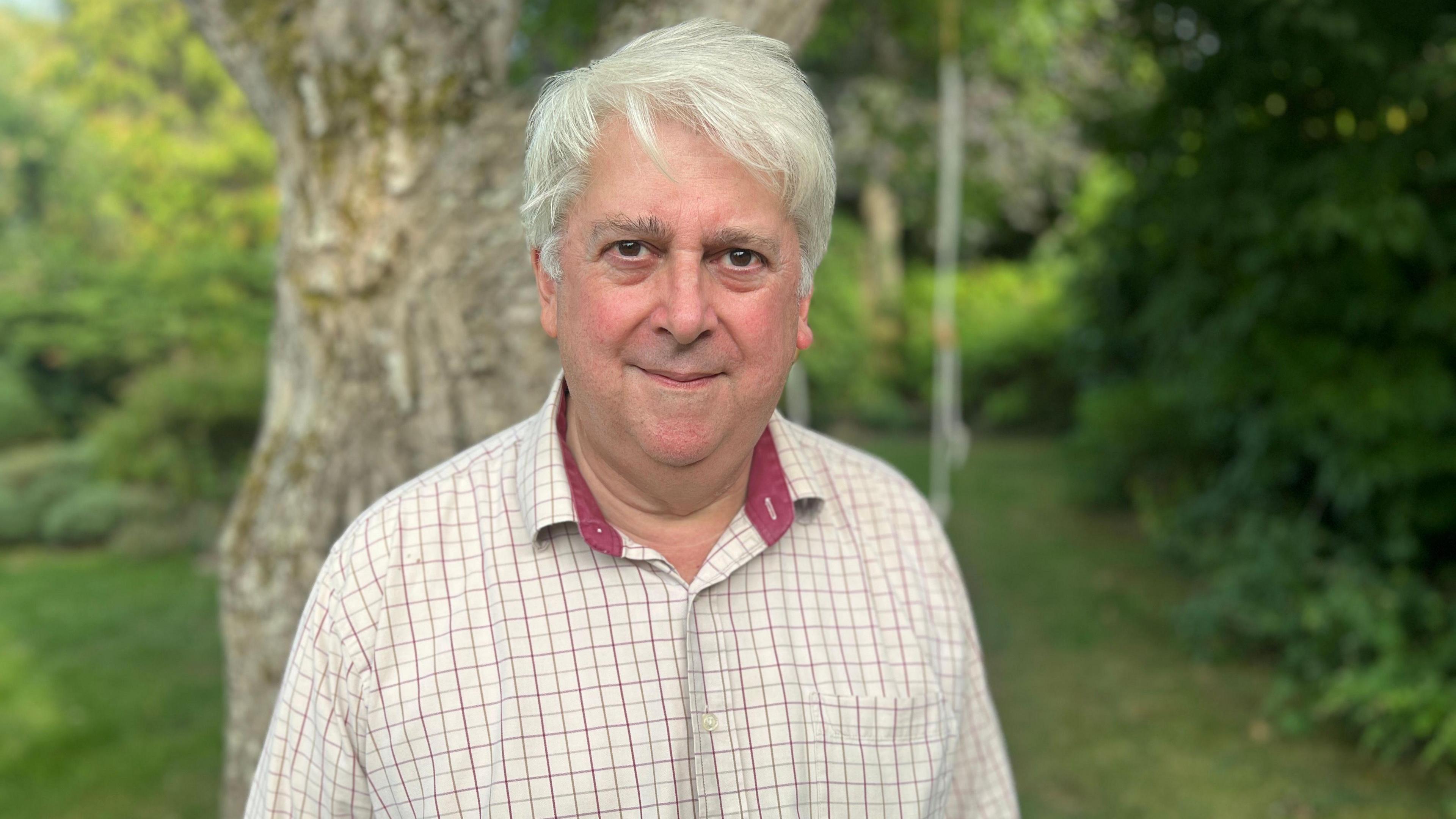 Headshot of James Matheson in his garden. Behind him is a tree which has a swing on it. James has white hair and is wearing a burgundy and white chequered shirt.