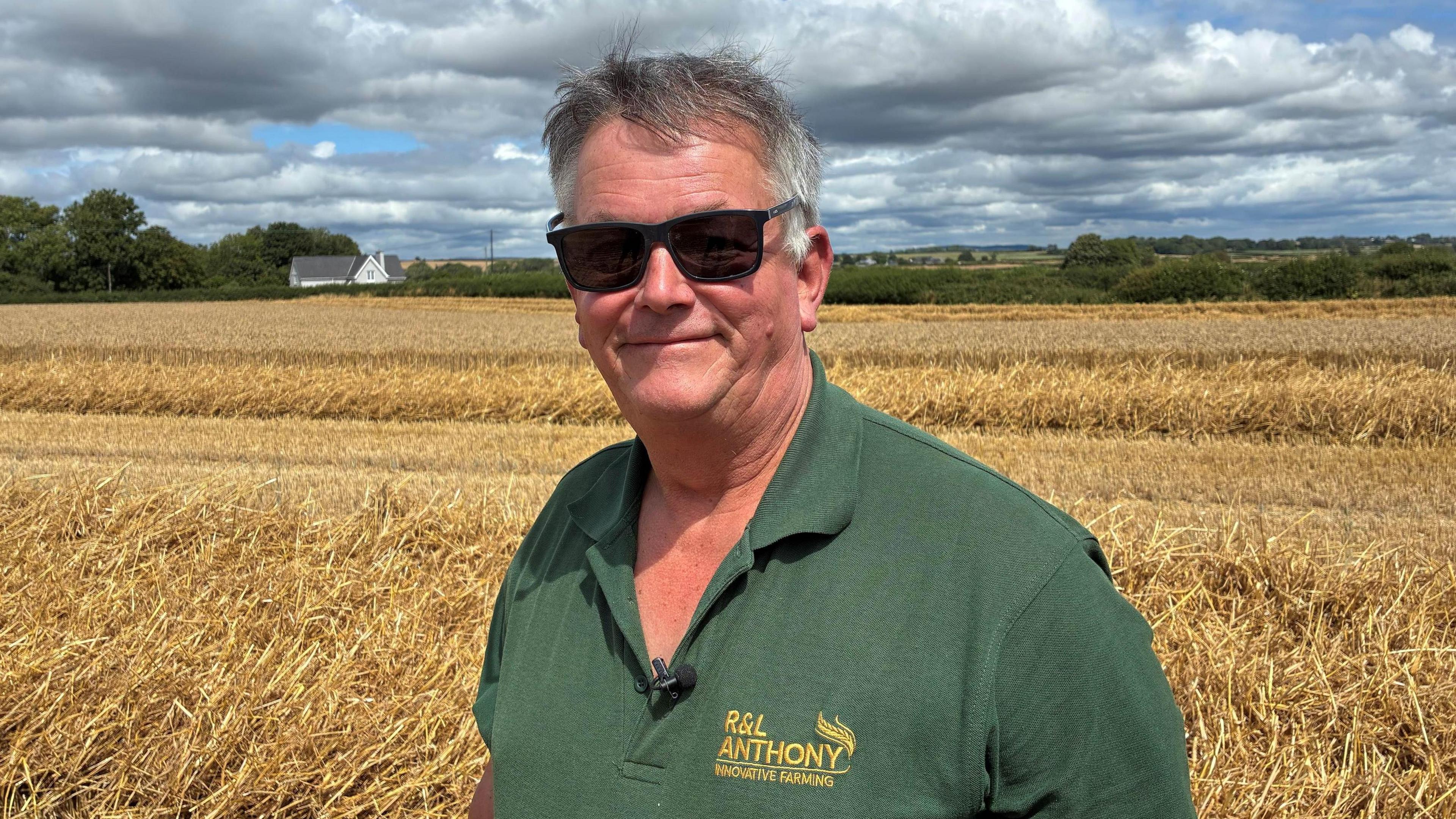 Richard Anthony is stood in front of a dry field on his farm. He is wearing a green polo top and sqaure frame sunglasses. He is smiling and looking at the camera.