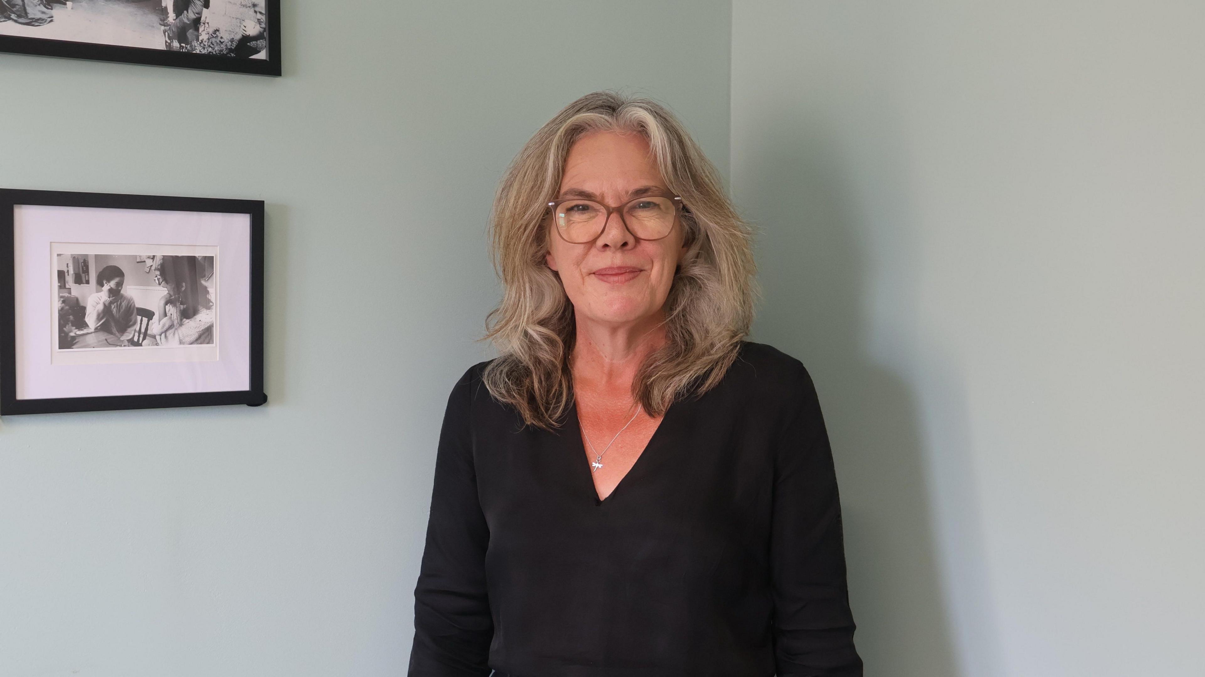 Tania is white and is wearing brown-framed glasses. She has fair hair and is wearing a black top with a silver necklace. She is standing in front of a wall and to her right is a framed black and white photograph of two women.