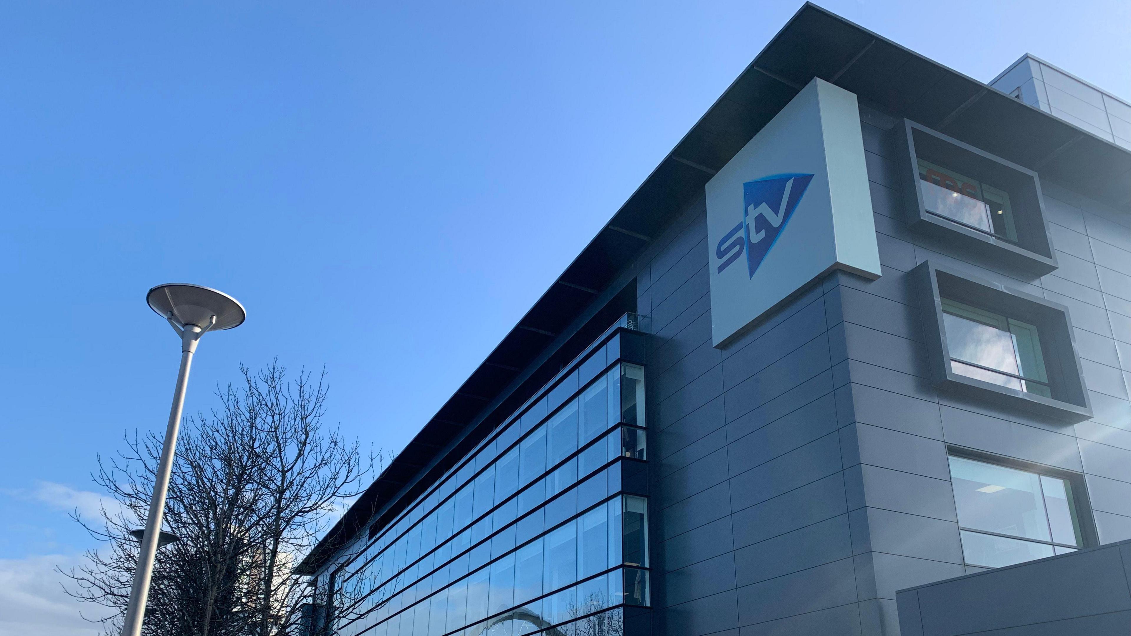 Exterior view of the STV building with modern glass and metal façade, featuring the STV logo on the corner. A row of leafless trees and a streetlamp line the pavement in front, with a clear blue sky overhead.