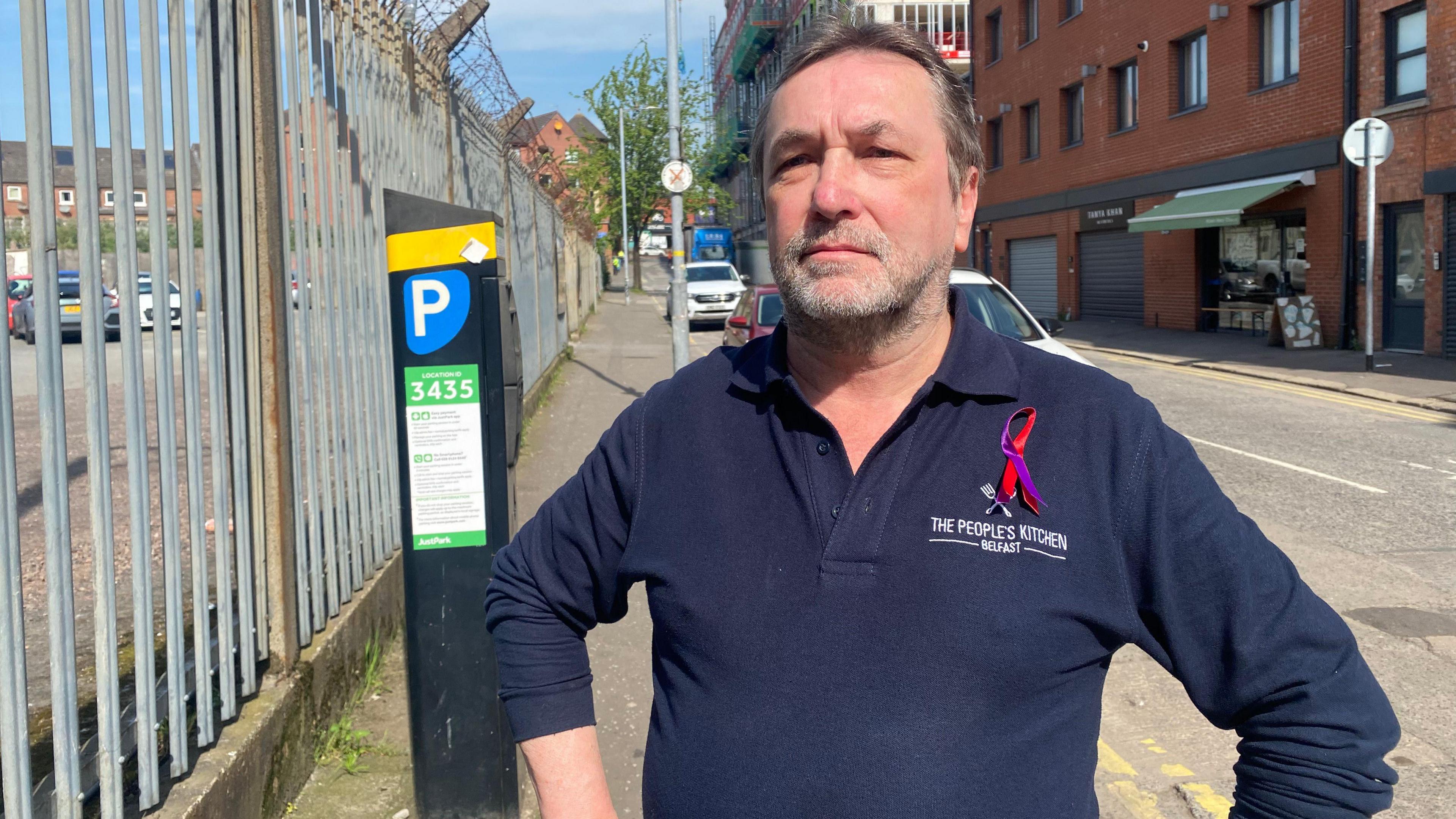 A man with grey longish hair and a beard wears a navy jumper, with The People's Kitchen, Belfast on the lapel. 