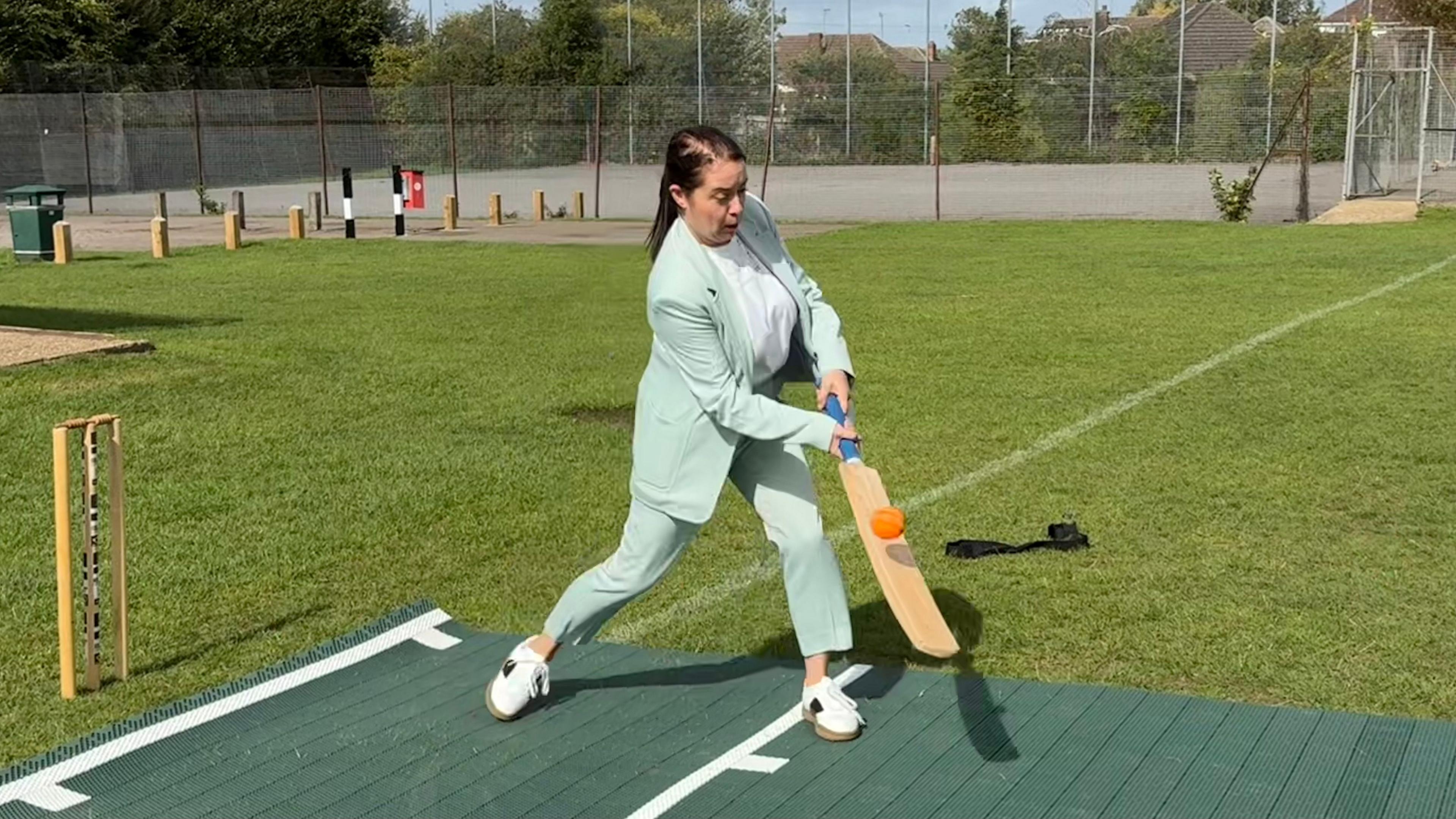 Stephanie Peacock with long dark hair, wearing a light green jacket and trousers, white shoes and white top. She is holding a bat which is just about to strike an airborne orange ball. She is on a green fabric square with wickets on the left. There are cricket nets in the background.