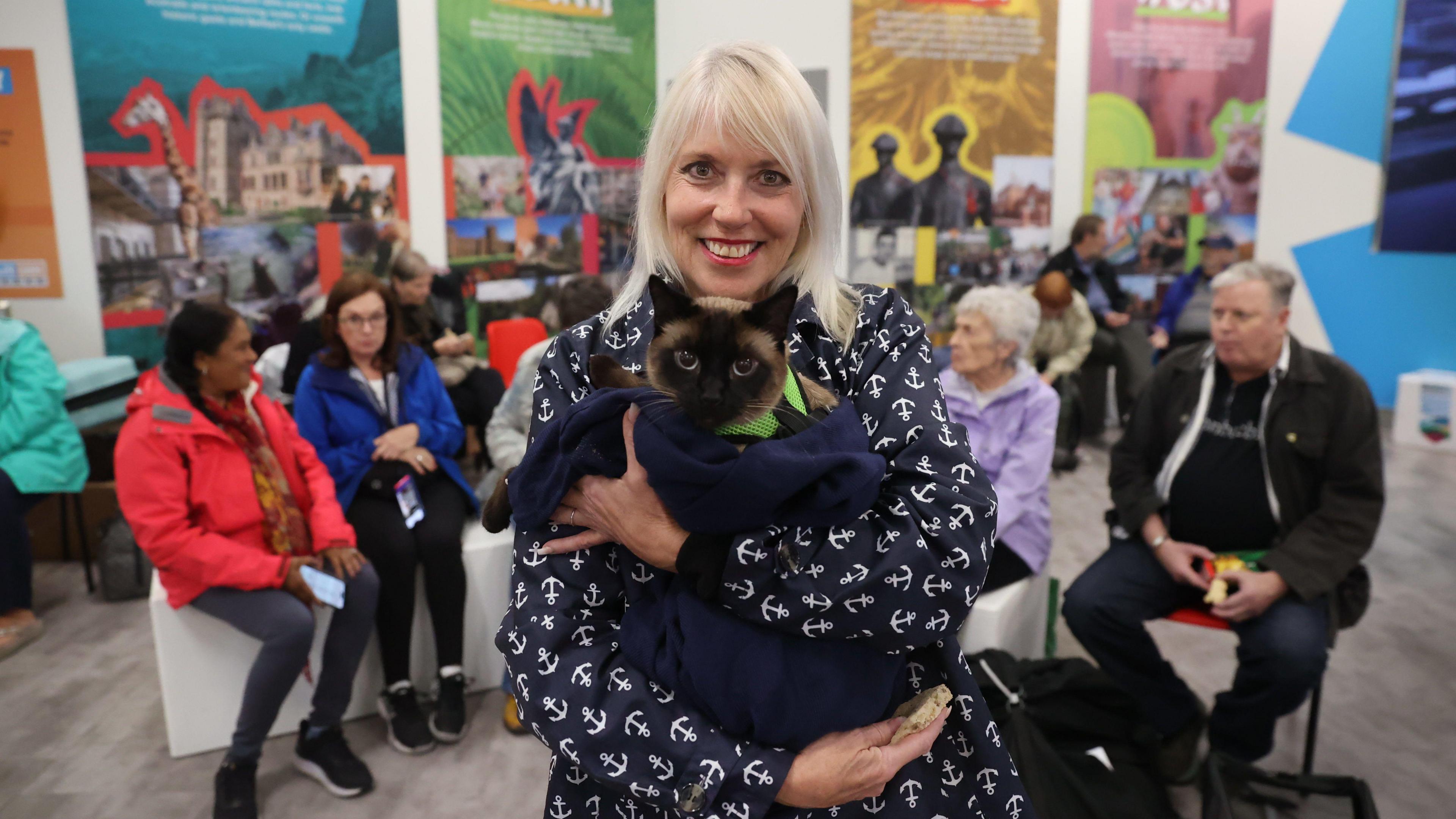 A woman, with blonde hair and wearing a navy jacket with white anchors on it, looking at the camera and smiling. She is holding a Siamese cat. There are people sitting down on chairs behind her. 