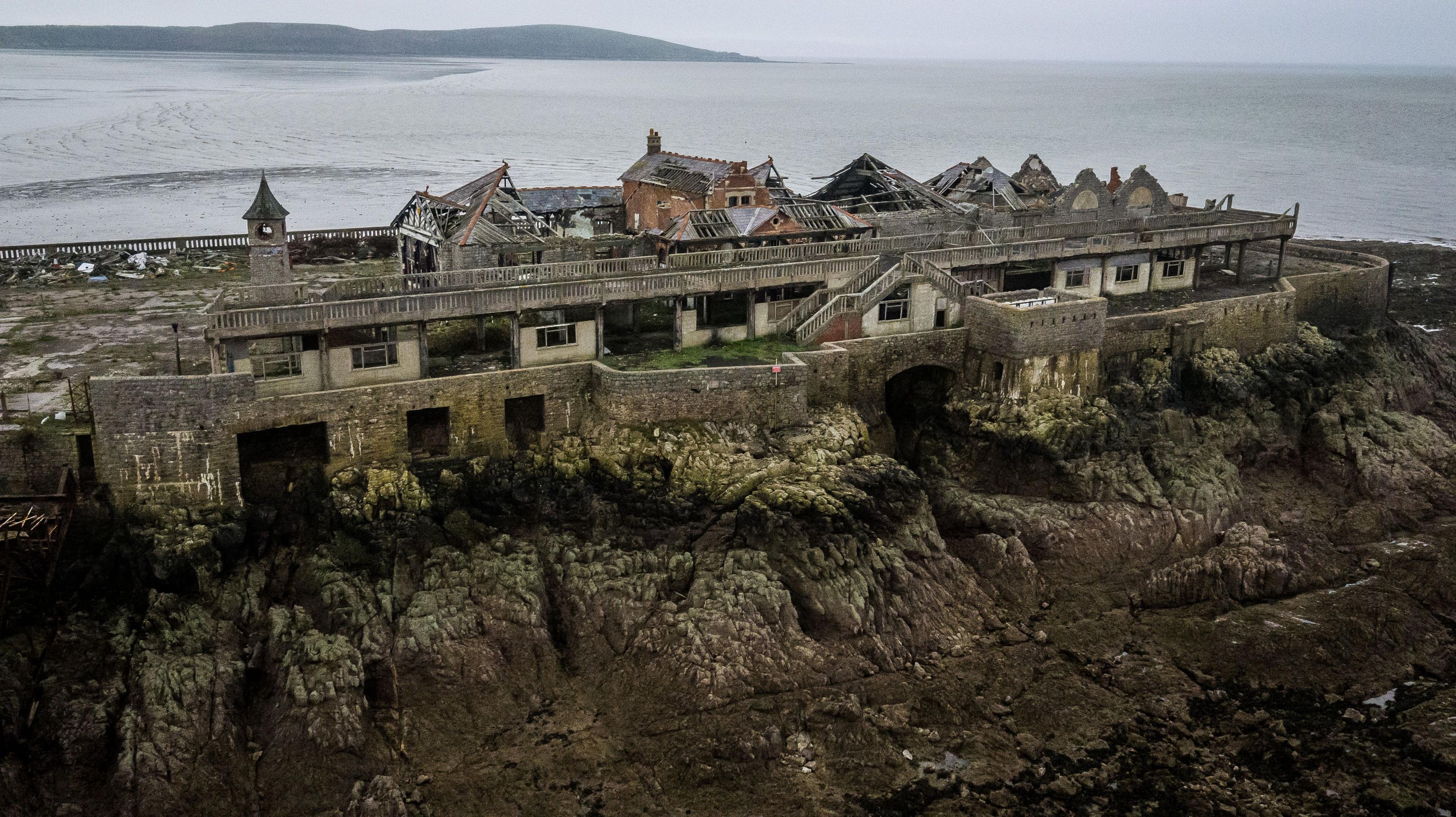 A dilapidated pier built on top of a rocky island. The majority of the building's roof has fallen in, with large sections of the building's walls damaged. The sea can be seen behind the pier, with the cloudy weather giving the image a dark and shadowy quality.