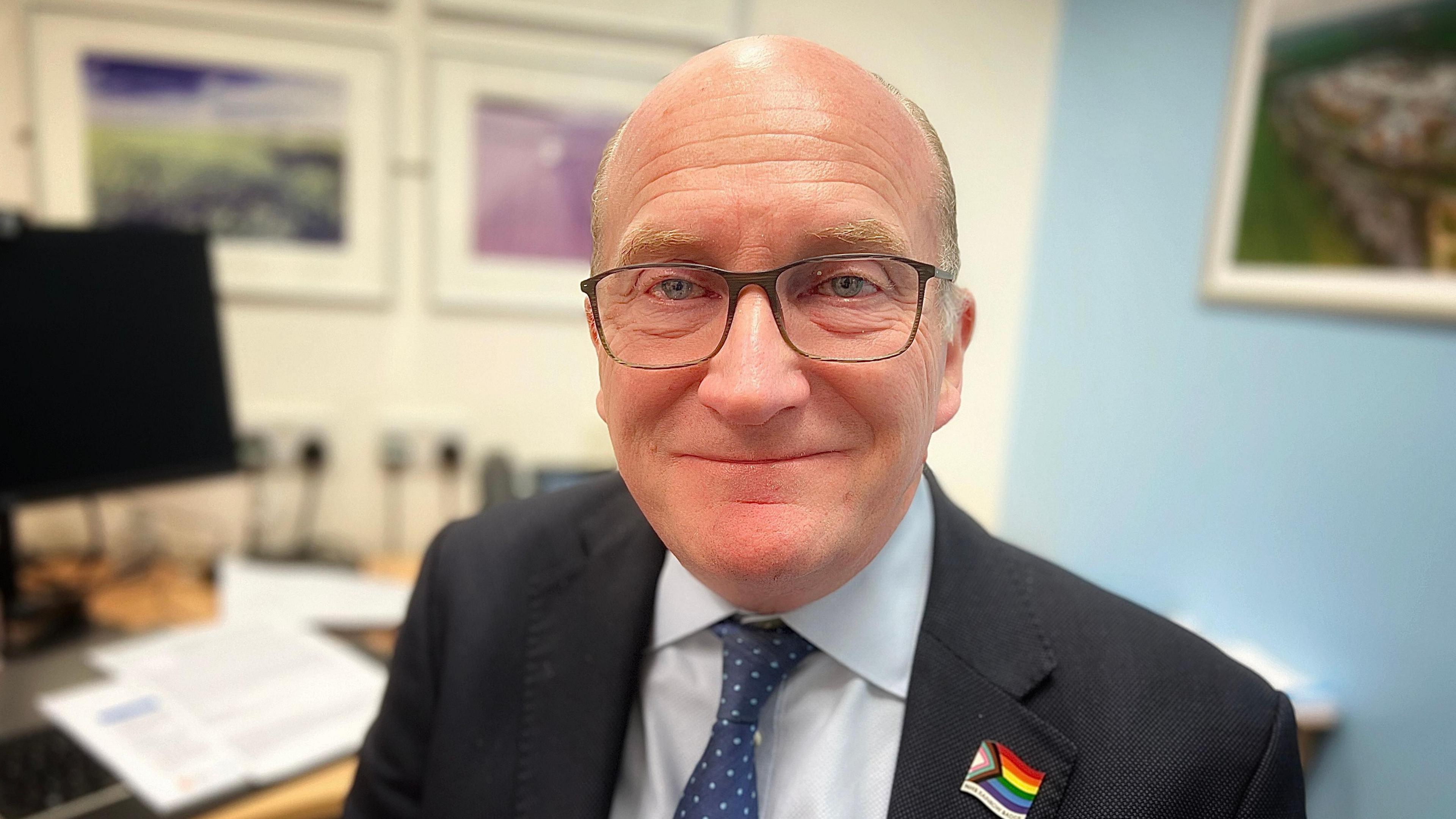 Nick Hulme with very short grey hair and glasses. He is smiling at the camera wearing a dark jacket, pale blue shirt and blue tie. There is a multicoloured flag emblem on his left shoulder. He is standing in an office with pictures on the wall and a large wooden desk on which a monitor is sitting.