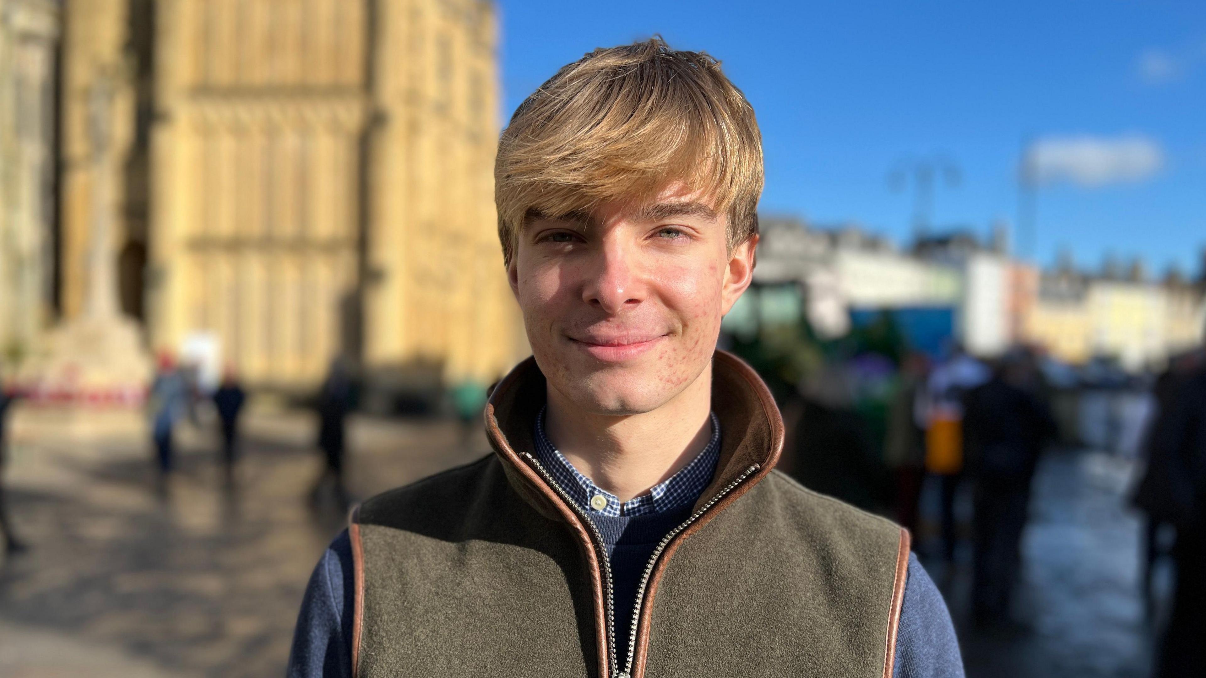 A young man with dirty blond hair stands in Cirencester town centre on a winter's day with the church behind him. He is wearing a long sleeved navy jumper and a brown fleece gilet.