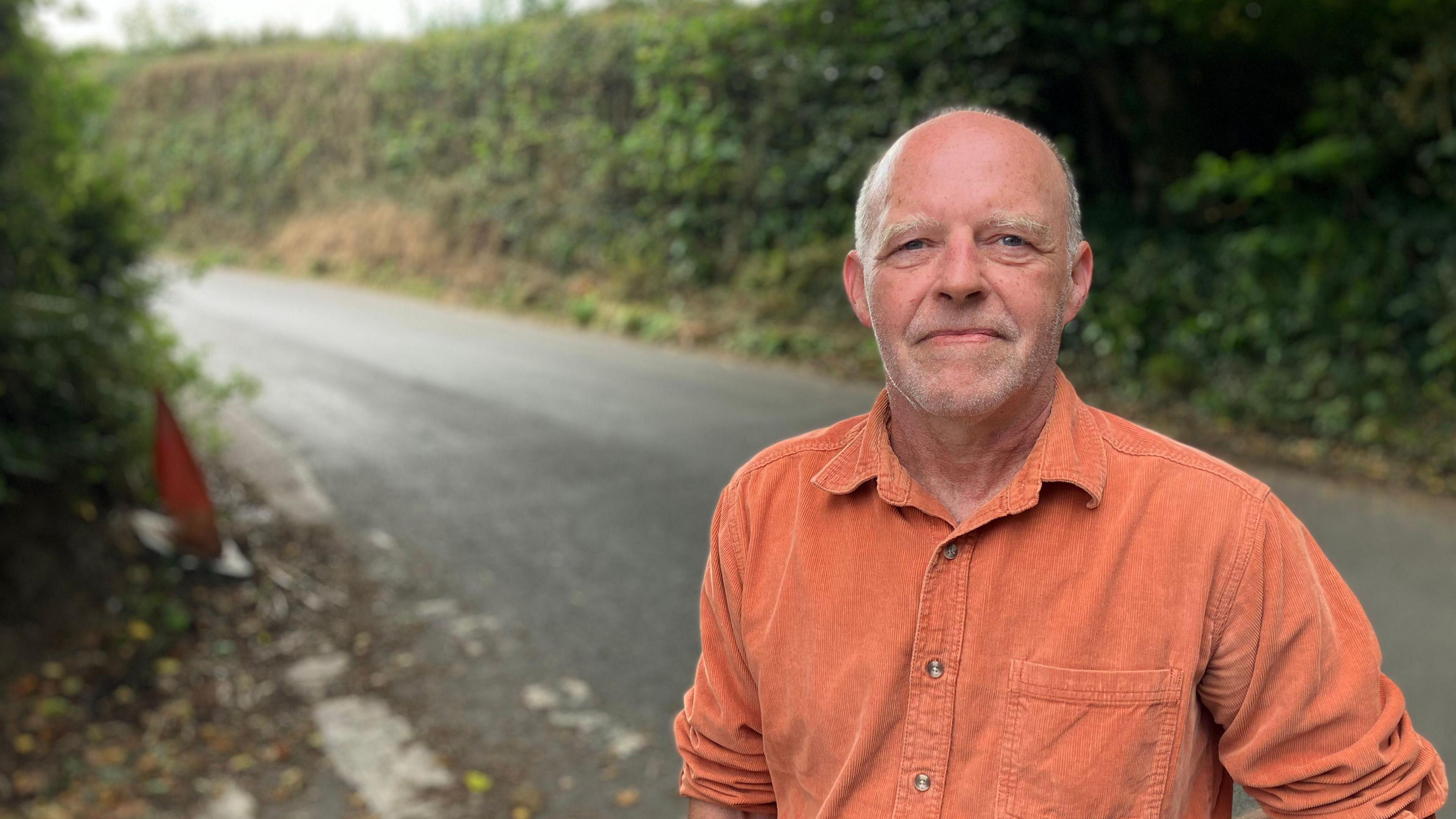 A man in an orange shirt stands at the side of a country lane, there are high Devon banks in the background, along with high hedgerows. The sky is overcast. 