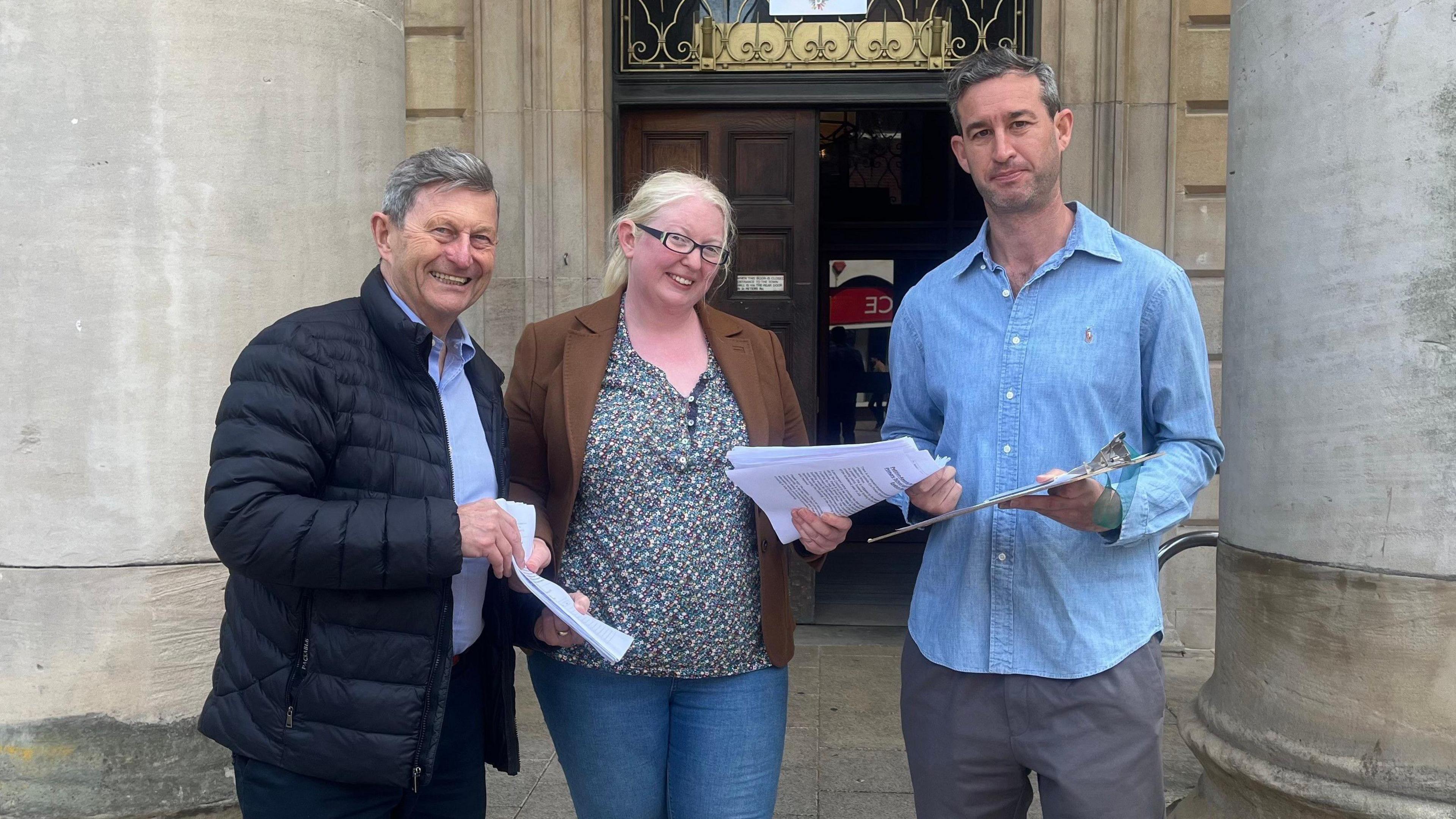 A woman is standing outside a large stone building between two men. She has blonde hair and glasses and is wearing a colourful top, a jacket and jeans. She is holding a pile of paper in her hands.