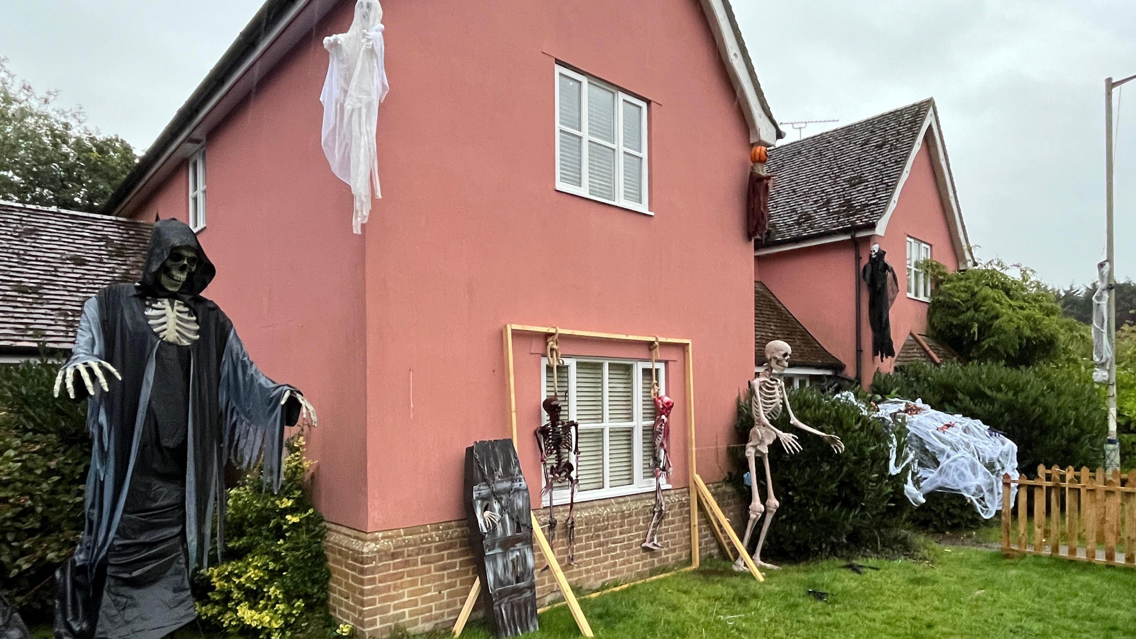 Another view of the front of the house with Halloween decorations on display outside of it. A large skeleton wearing a black reaper cape is on the left of the image. Ghost decorations hang from the top of the house. There is also a black coffin resting on the house. 
