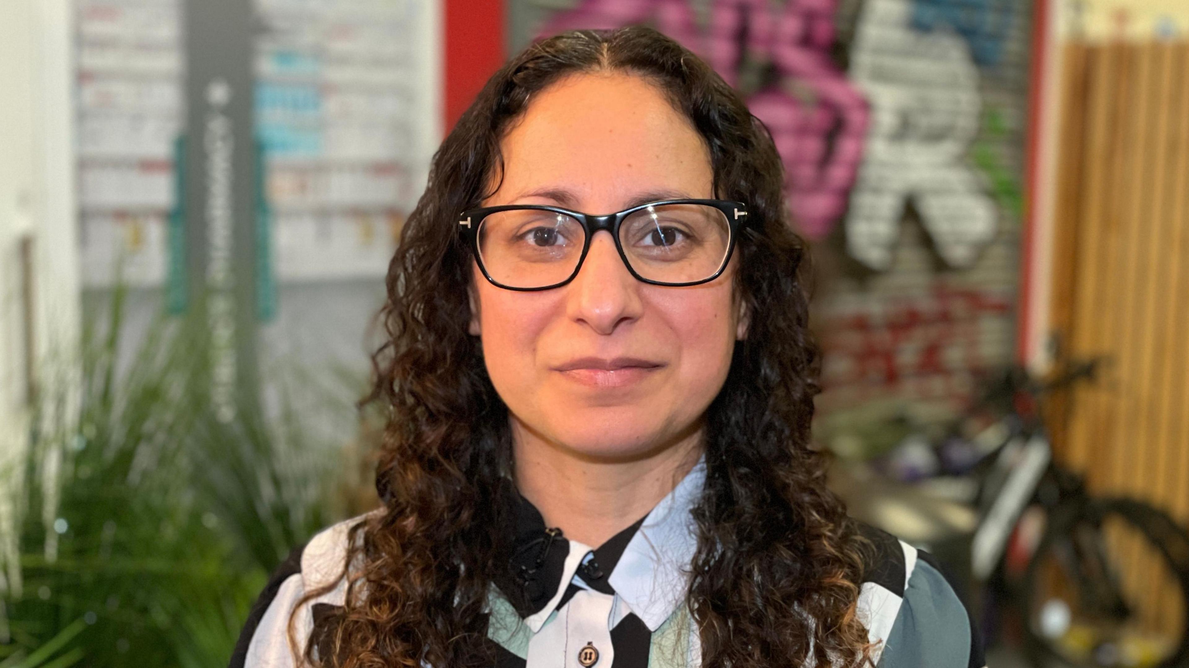 Maya Joseph-Hussain is the chief executive of the Safety Centre in Milton Keynes. She has long, dark brown hair in tight curls and is wearing a pair of black glasses. She is looking directly at the camera. Behind her, out of focus, is a bush, a bicycle and a wall covered in graffiti. This photograph was taken on a fake street built inside the Safety Centre to teach children about hazards.