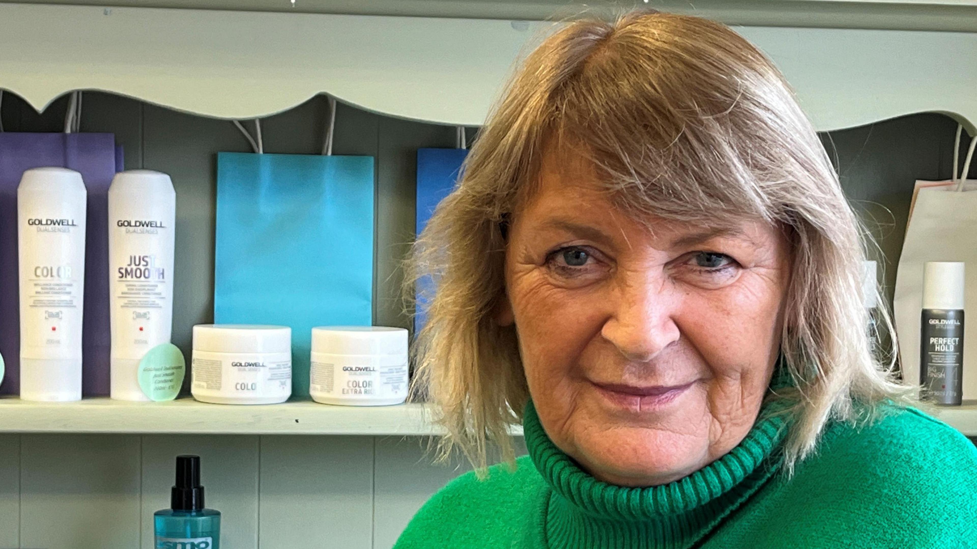 Pauline Liberti - head and shoulders image of her wearing a green, polo neck jumper.  She has shoulder length, fair hair. In the background is a pale green shelf with several hair products on display.