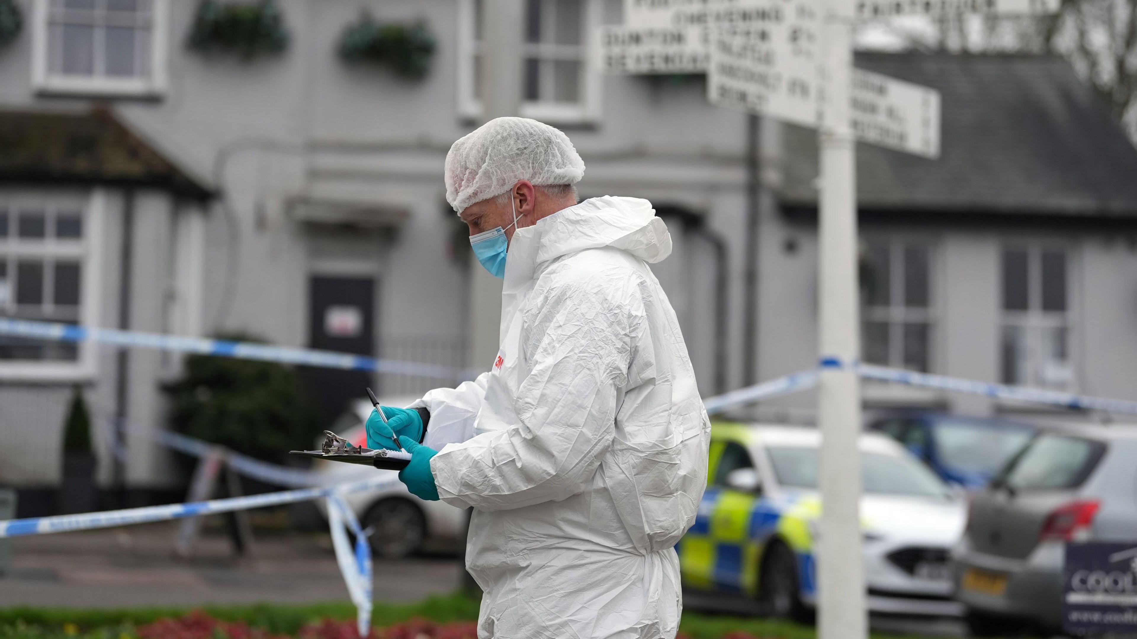 A police forensic officer writes on a clipboard, wearing a white gown and blue plastic gloves. 