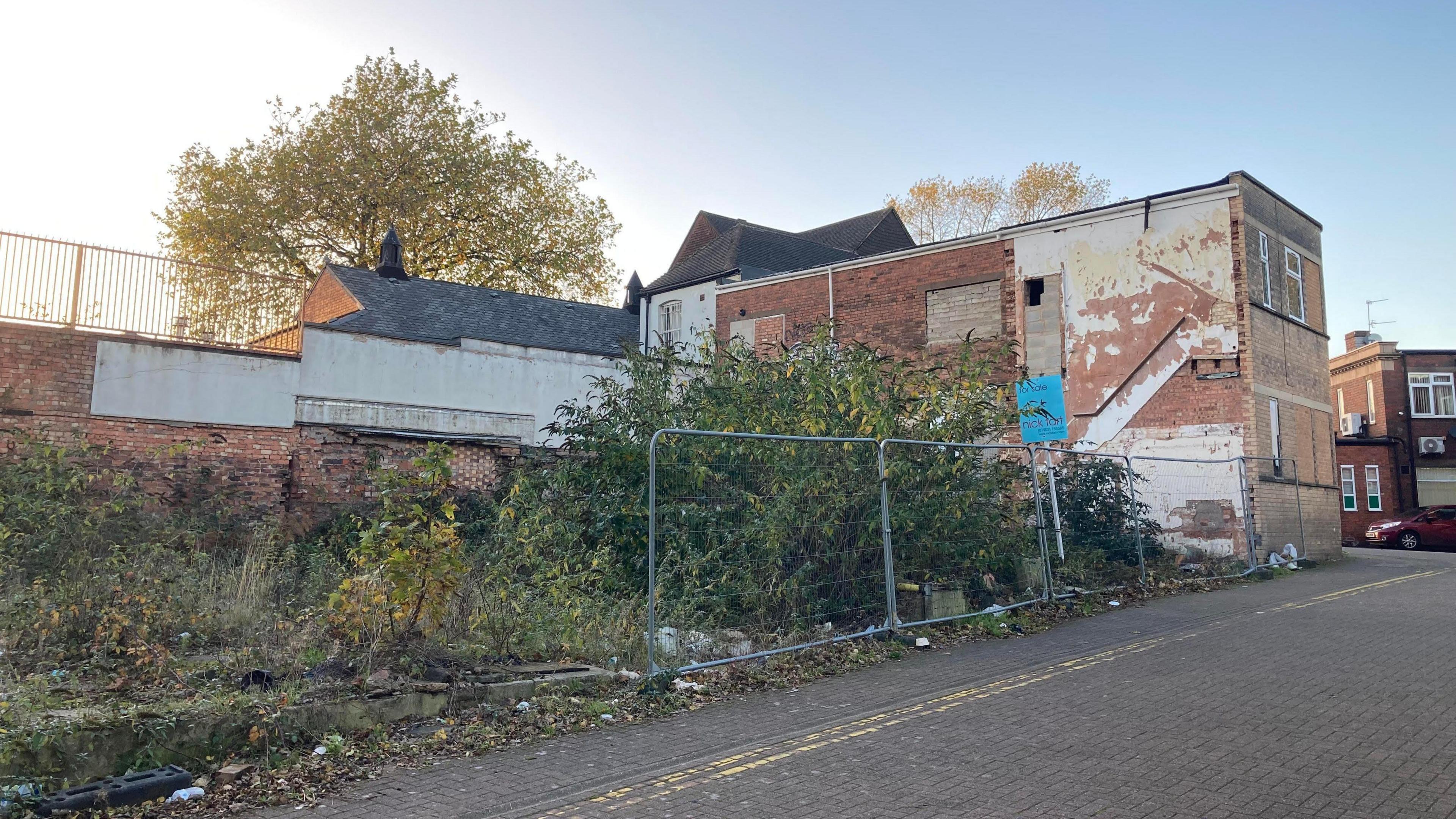 An overgrown plot of land next to a road and a derelict red brick building. There's metal fencing in front of part of the land. At the back is a red brick wall with fencing on top and a tree rising avove it.
