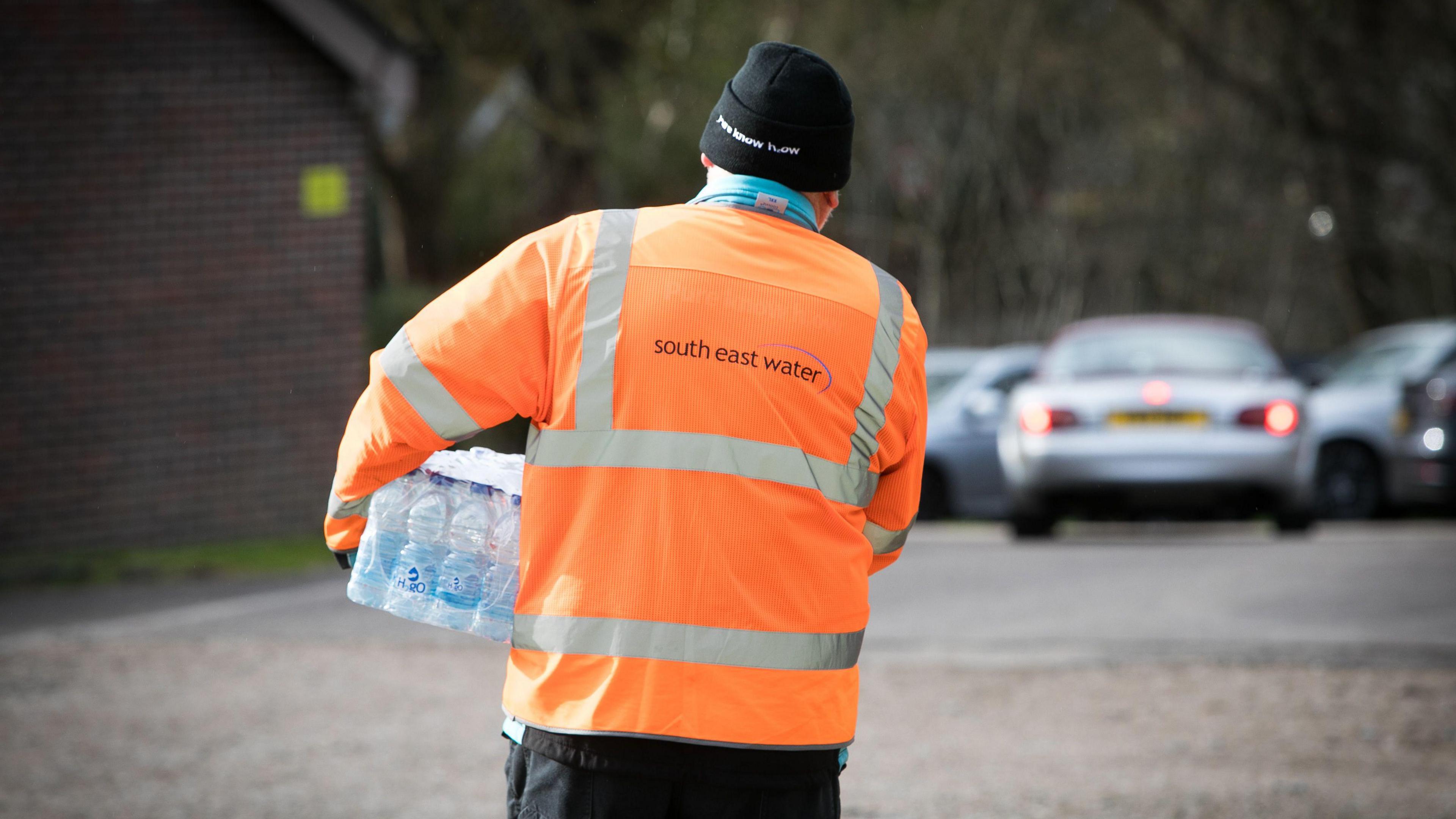 A man in a high-visibility jacket walking away from the camera while holding a packet of water bottles. He is wearing a black hat. There are cars in the background of what appears to be a car park.