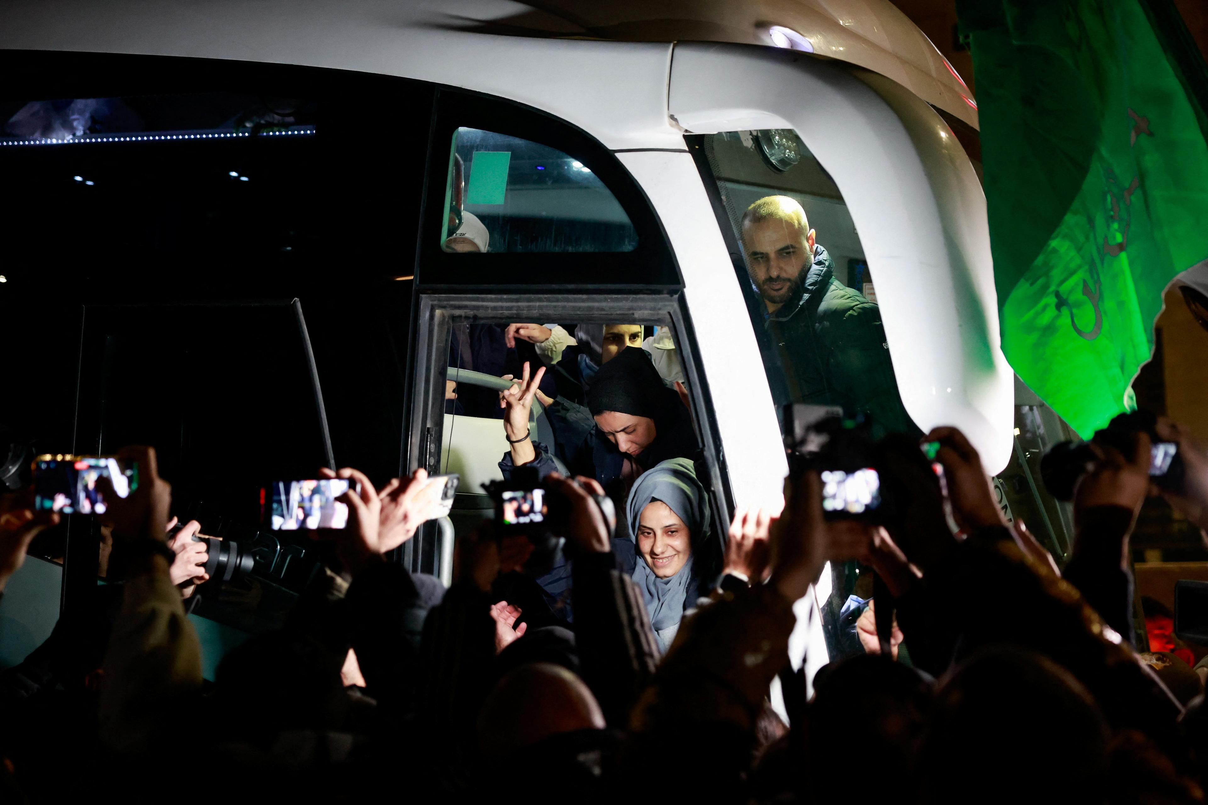 A smiling woman gets off a coach and is met by a cheering crowd, with many people holding smartphones in the air