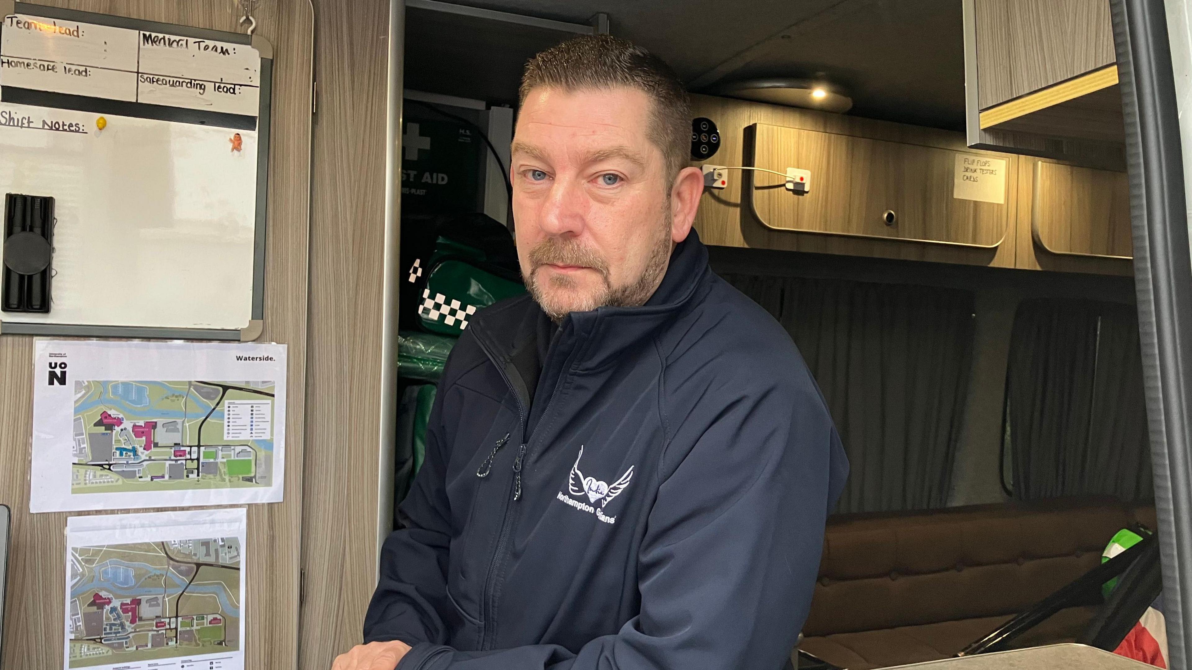 Man wears a navy jacket and is in the back of a van with information displayed on the side behind him