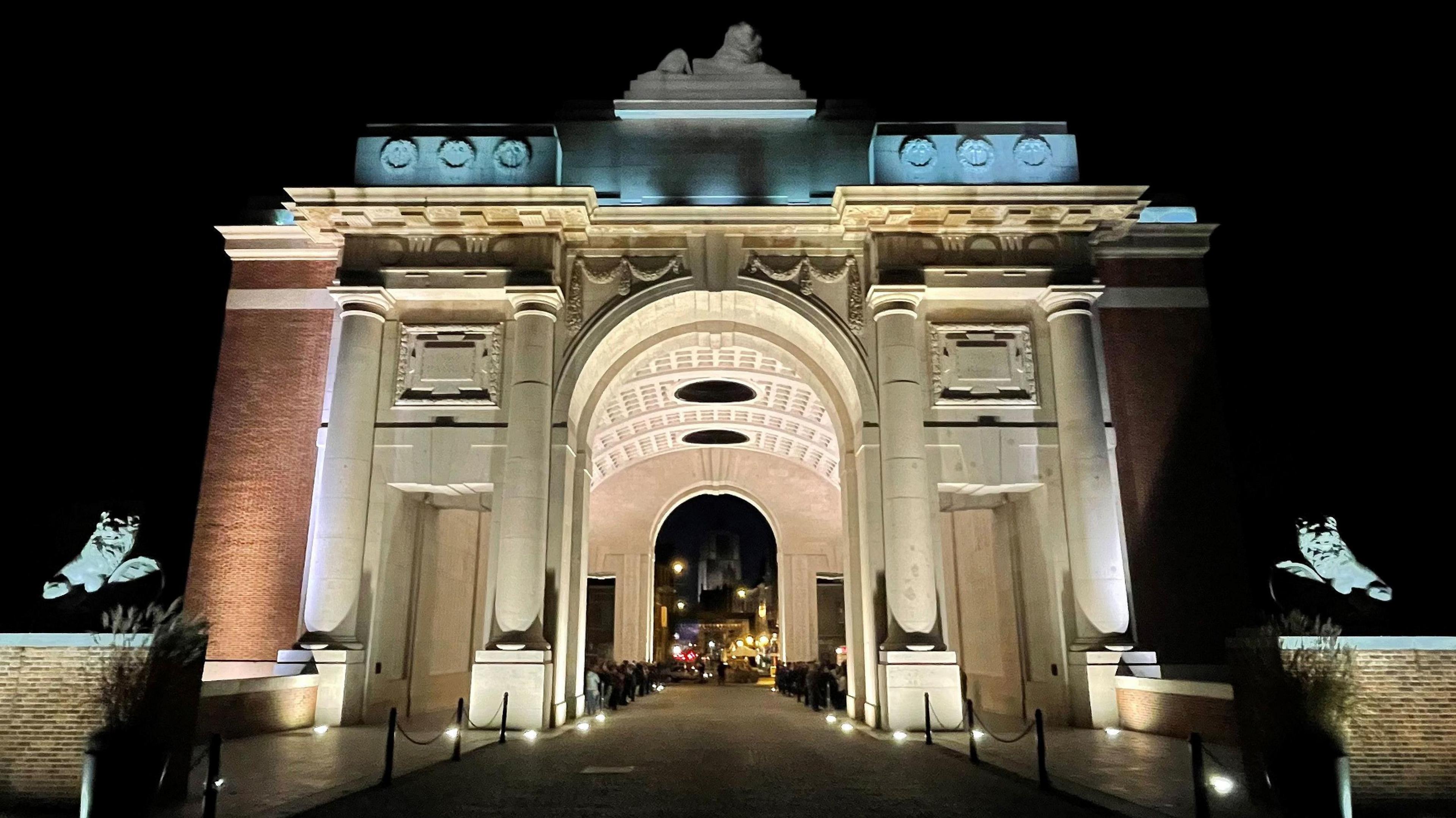 The Menin Gate is illuminated at night. It has tall marble columns either side of an archway that is the entrance to a large tunnel running through the structure. A pathway leading to the gate is lit up by spotlights.