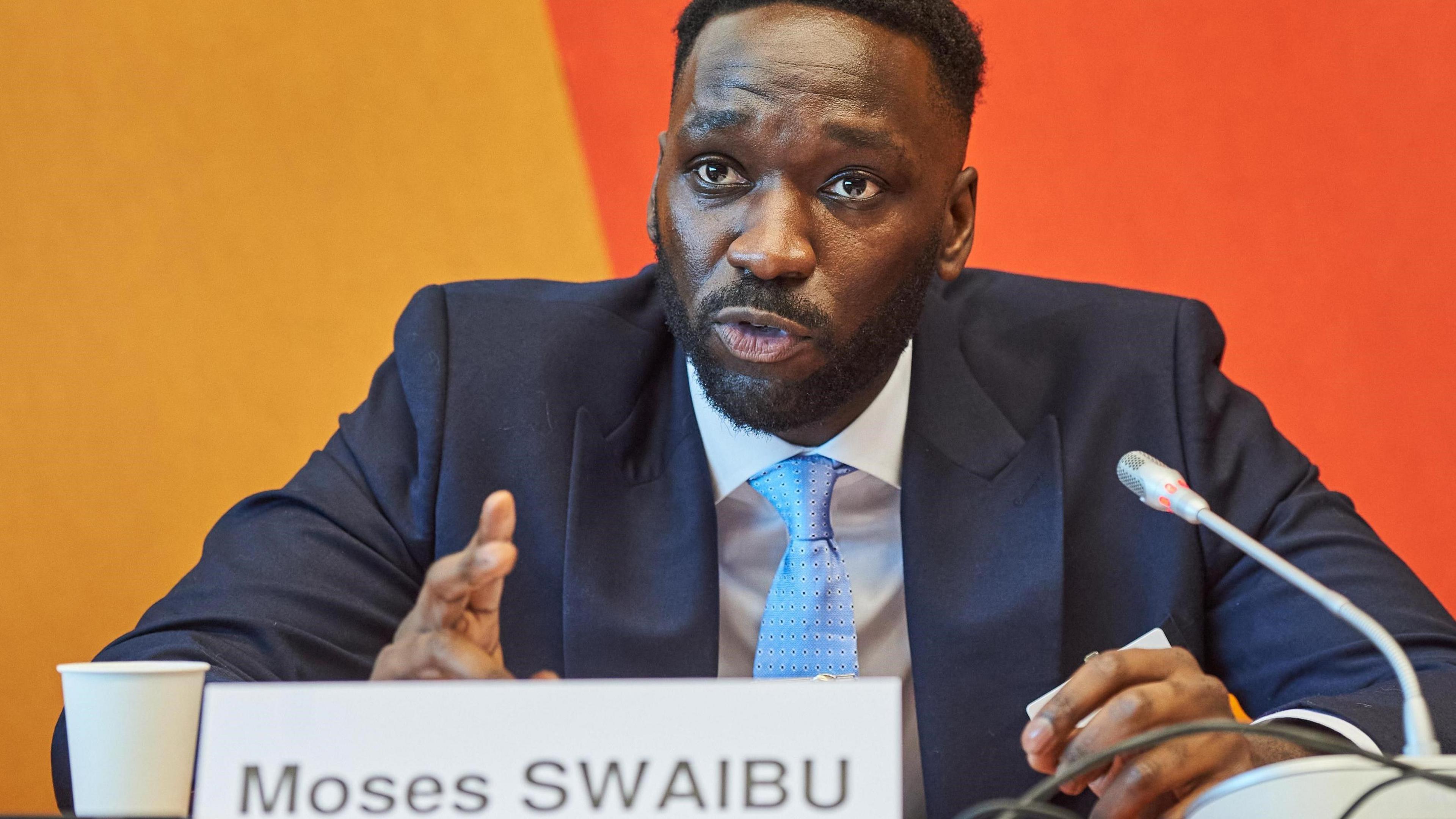 Moses Swaibu speaks at a conference, weaing a suit with a name plate bearing his name in front of him