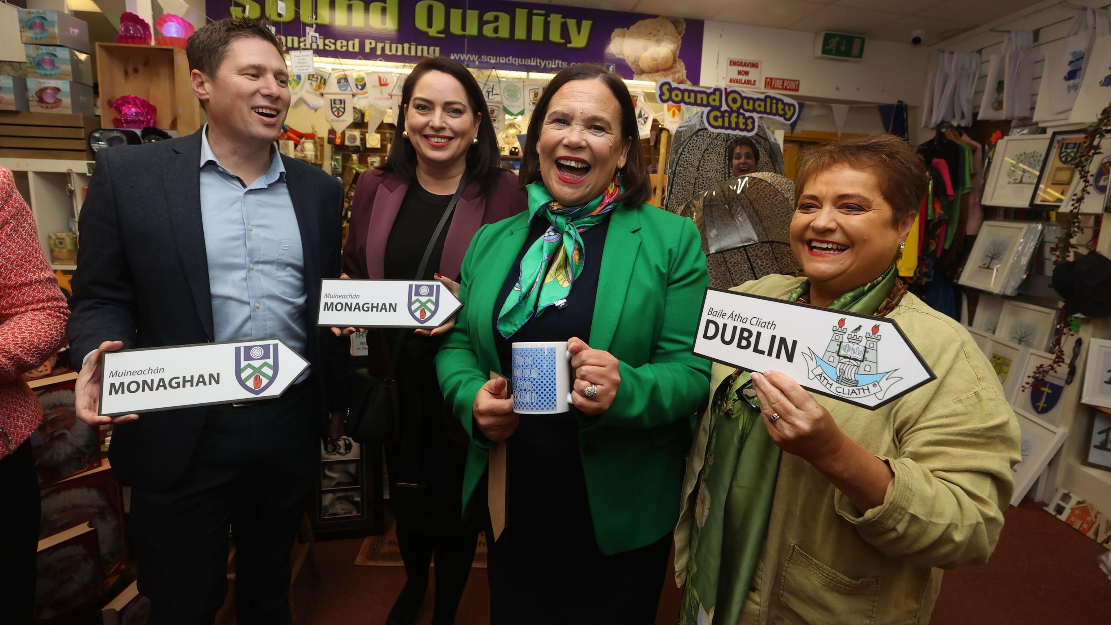 From left to right, there is a man wearing a navy suit and blue shirt, a woman with a black dress and purple jacket, Mary Lou McDonald, wearing a black dress and green jacket and a woman wearing a cream shirt. They are all smiling at the camera. They are standing in a gift shop. 