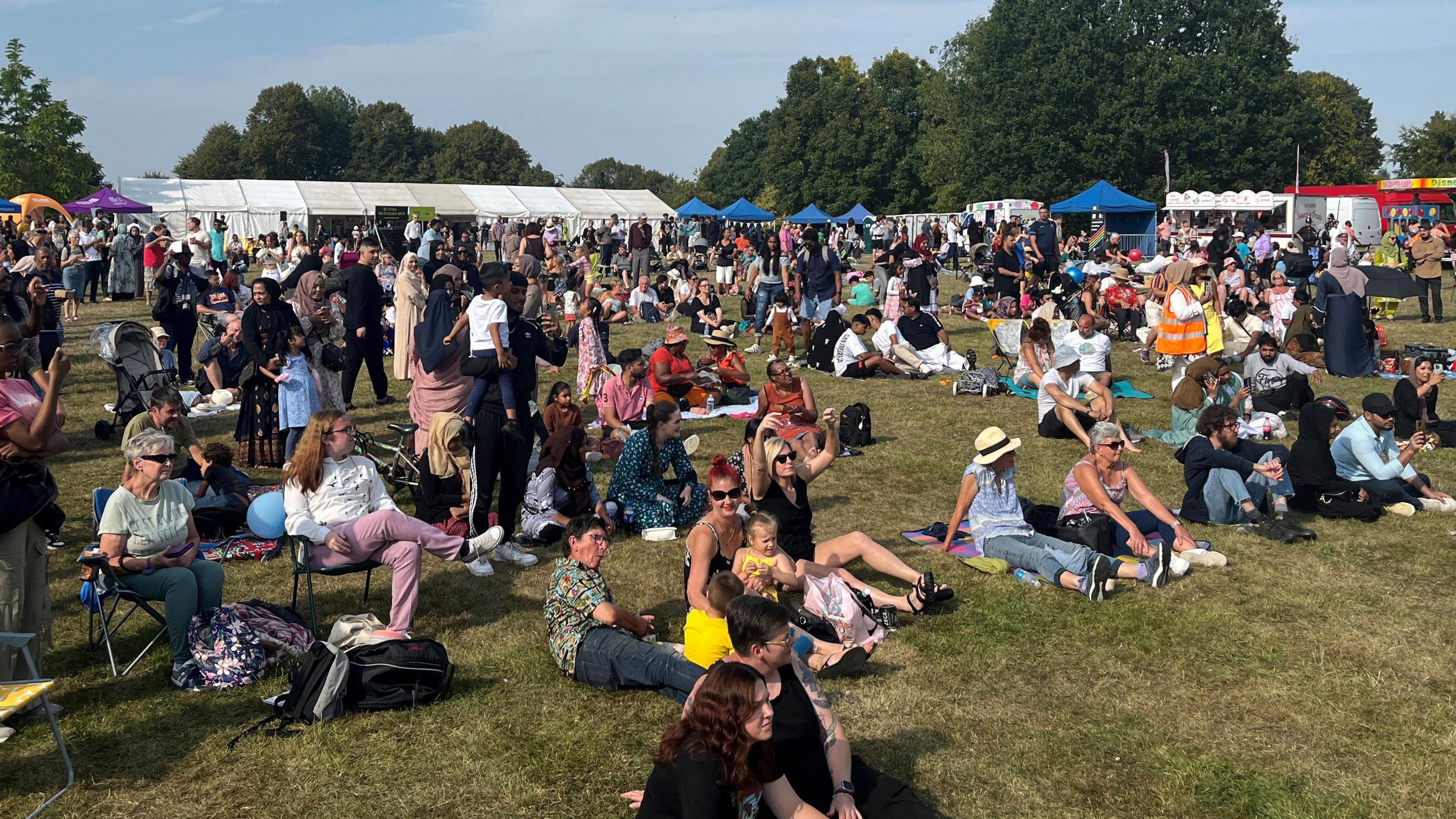 A crowd of people, of various nationalities, sat or standing on the grass in a park, with marquees behind them