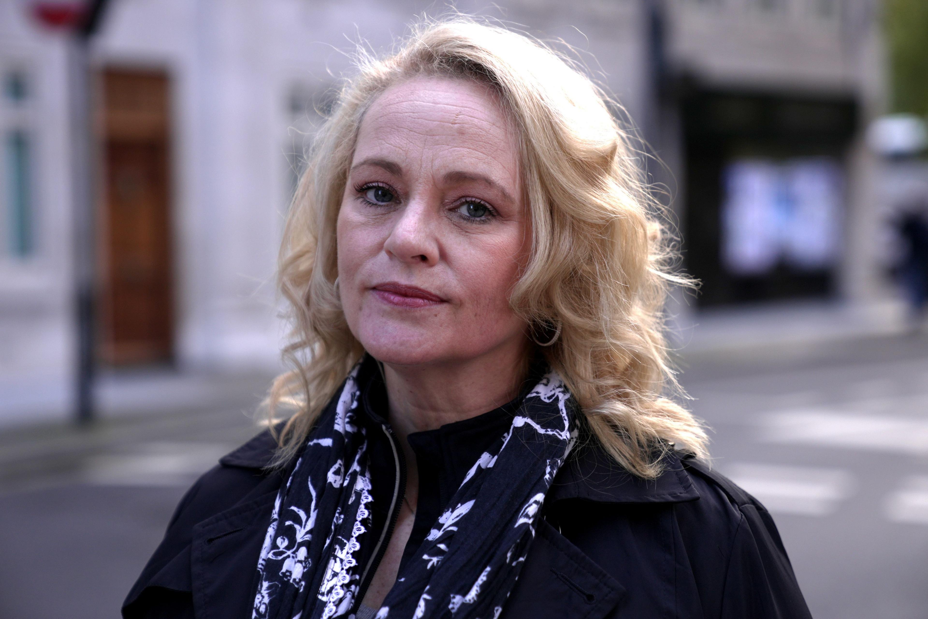 Amy looking directly at the camera, wearing a serious expression. Sunlight catches on her blonde hair and she is wearing a black jacket and monochrome scarf. 