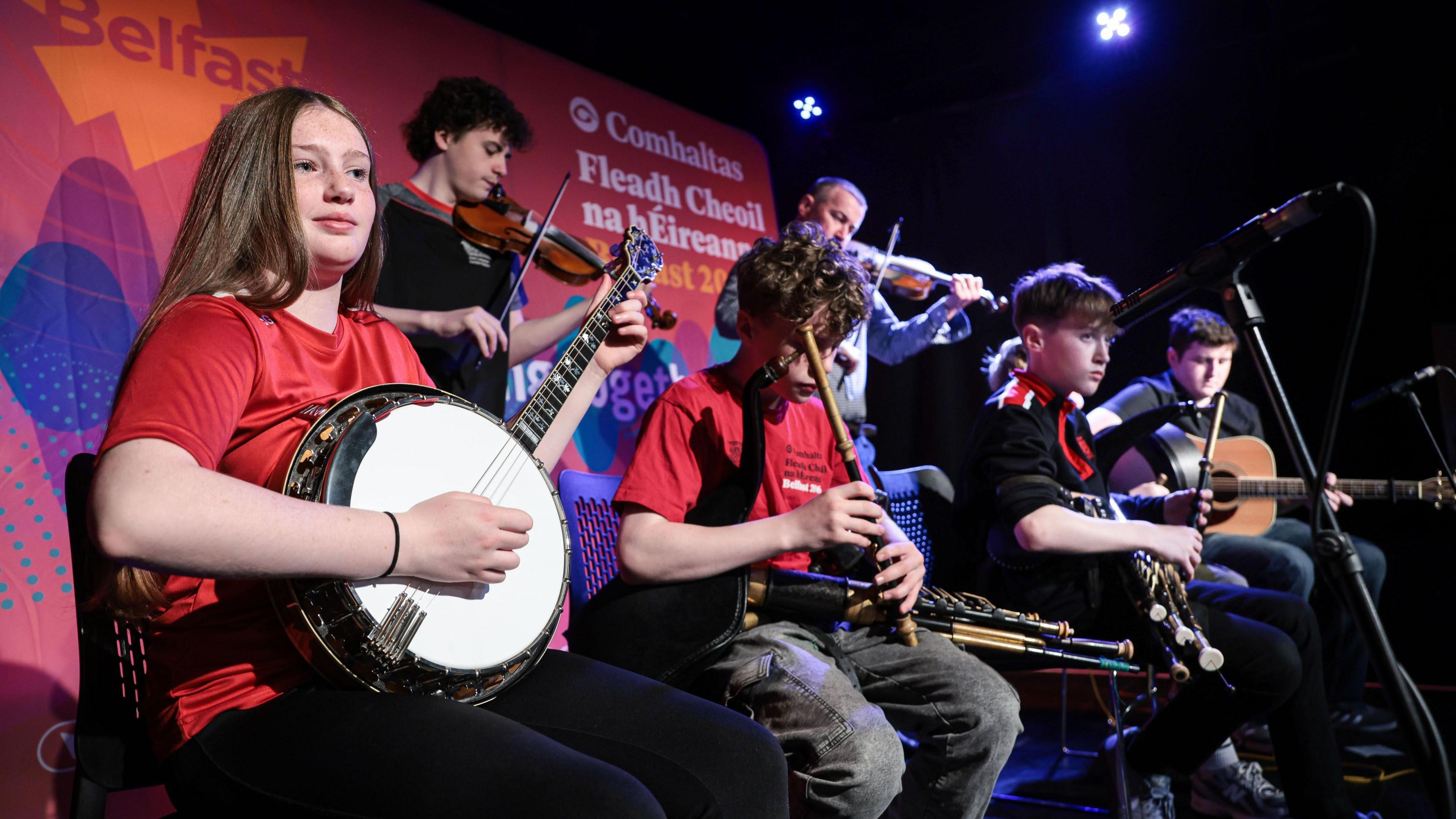 Six young people playing a mixture of instruments - in the foreground a girl with long brown hair, wearing a red tshirt, plays the banjo. Next to her is a boy in a red shirt and grey jeans, playing a flute. He has medium length brown hair, as do the other three boys - playing fiddle, guitar and pipes. The group are sat in front of a pink Belfast Fleadh backdrop. 