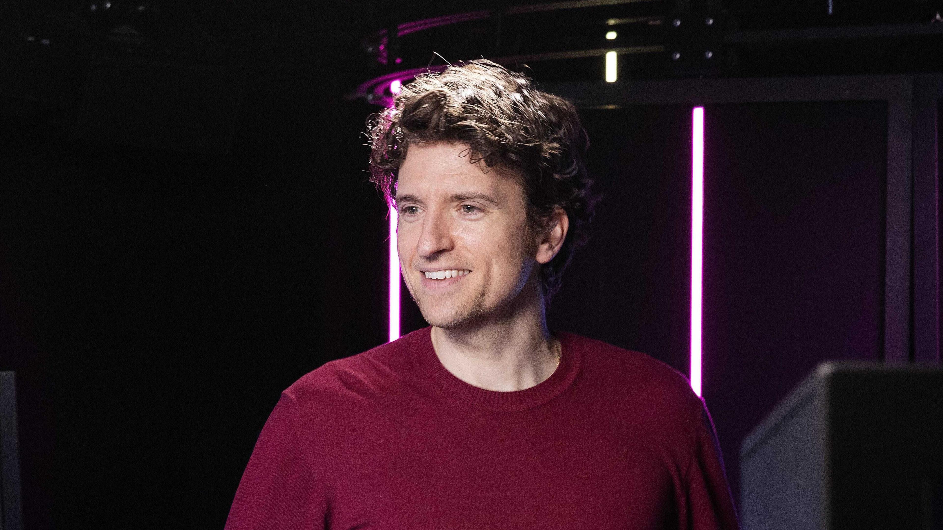 Greg James smiling left of the camera. He has curly hair and is wearing a maroon t-shirt. He's in a radio studio lit up with pink lights.