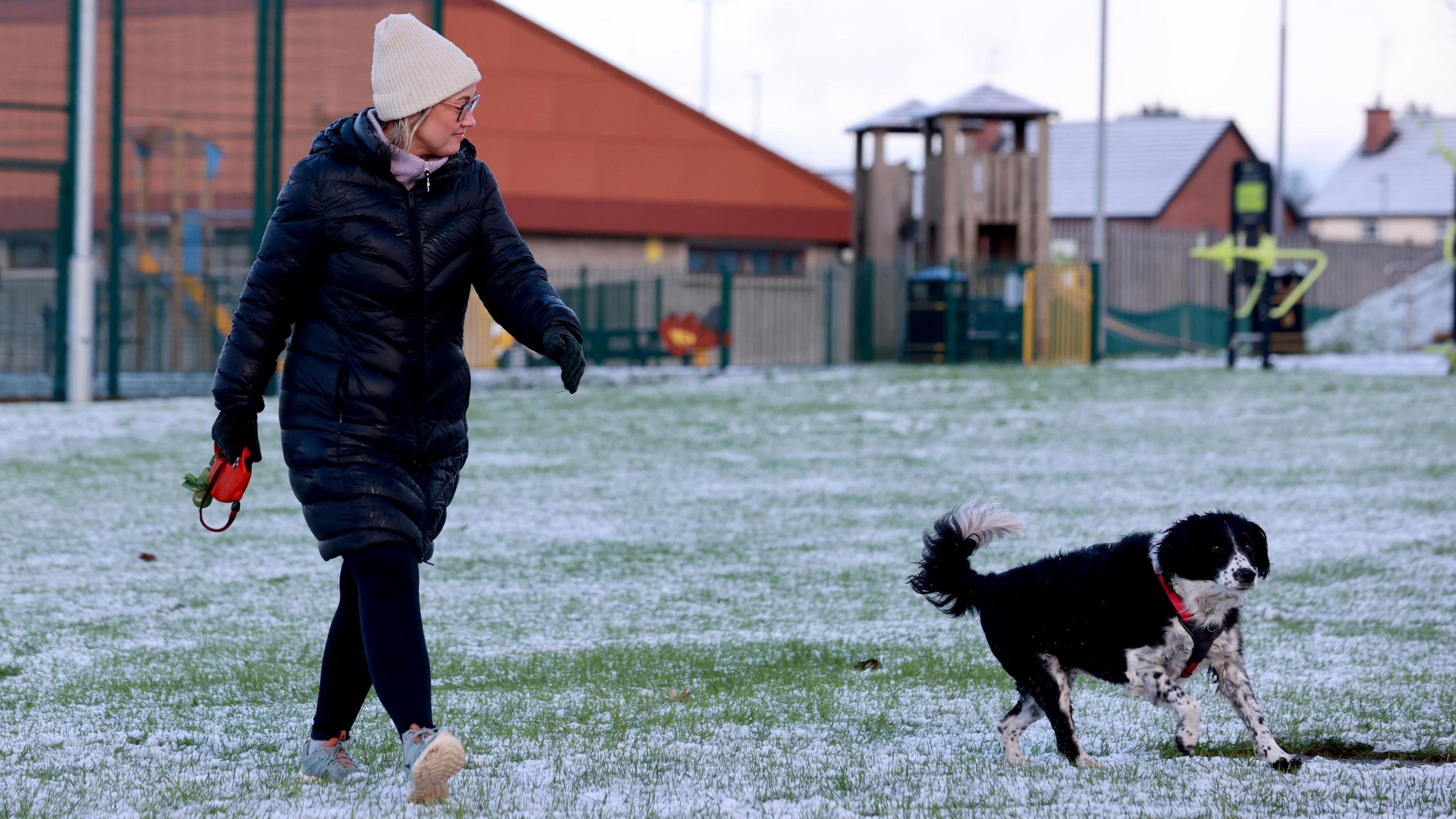 A woman is wearing a white knit hat, a long black puffer coat and black leggings and she walks her black and white dog Bonnie in a park in Portglenone. There are green gates in the background and in the foreground is a small amount of snow on the grass.