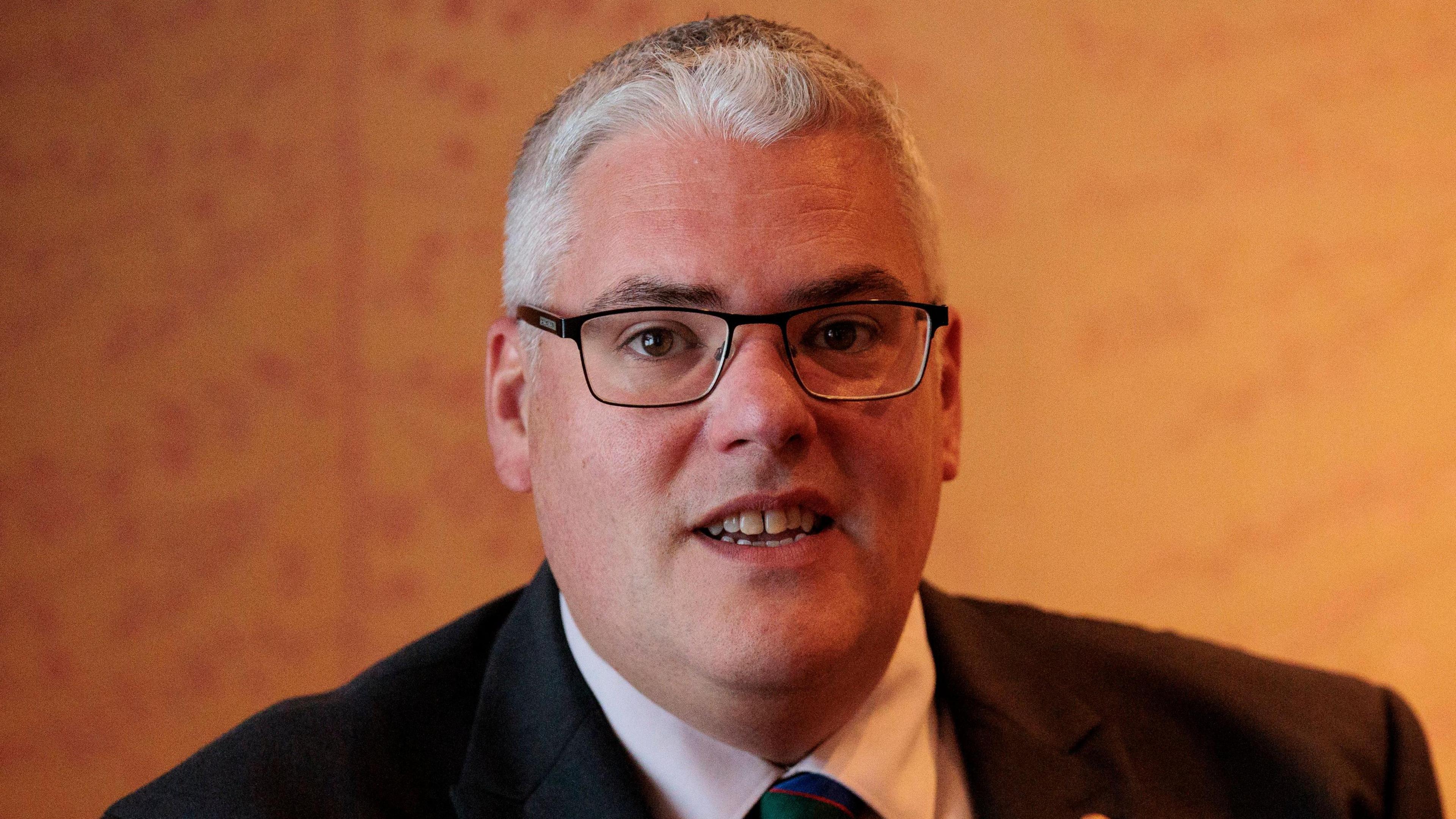 Gavin Robinson in front of an orange wall. He is wearing a suit and has grey hair.