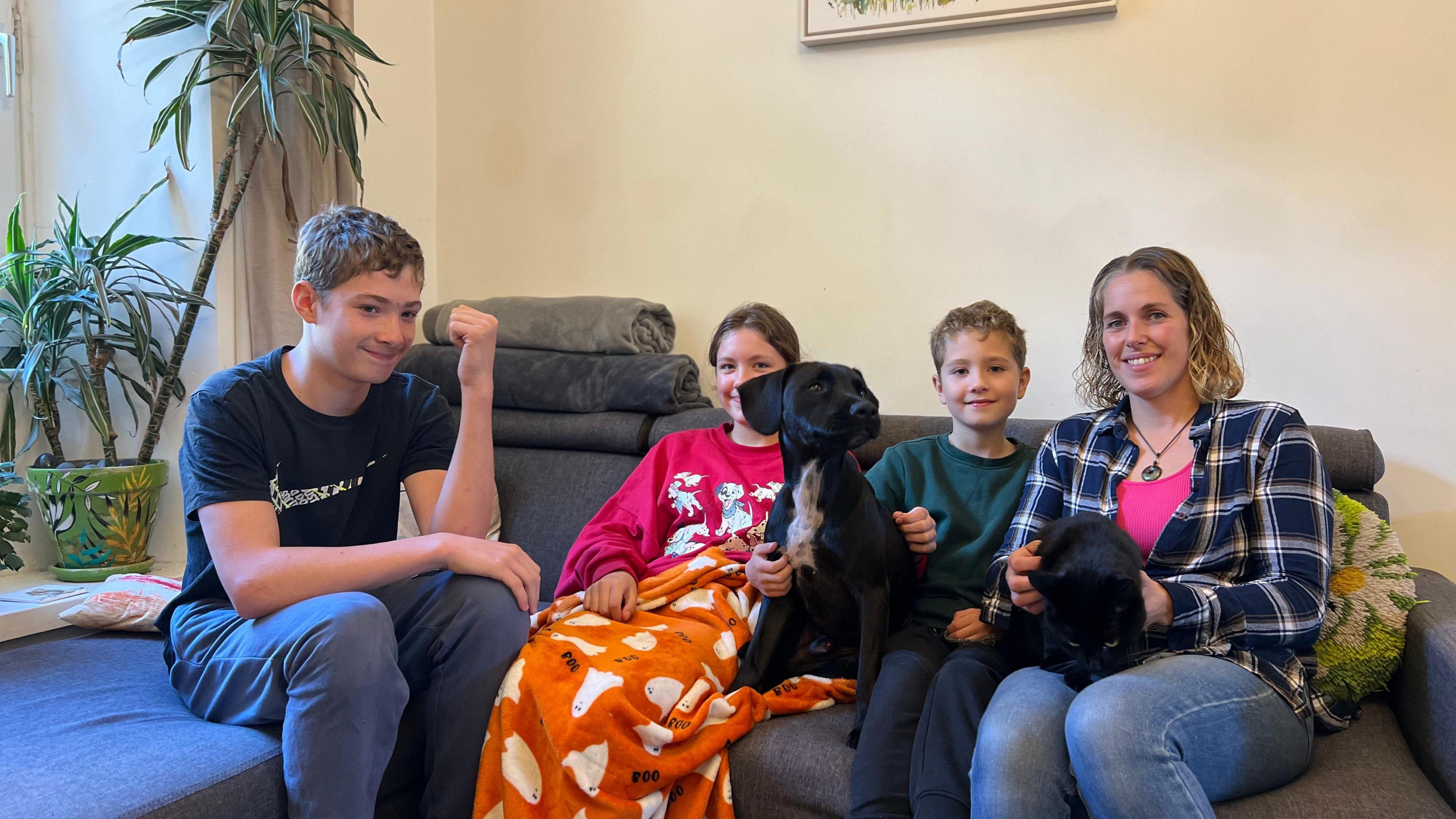 Connor, Keira, Reese and Hayley all sat on the sofa together. Hayley is holding Polo, a black cat, and there is a black dog sat to the right of Keira. They are all smiling at the camera. 