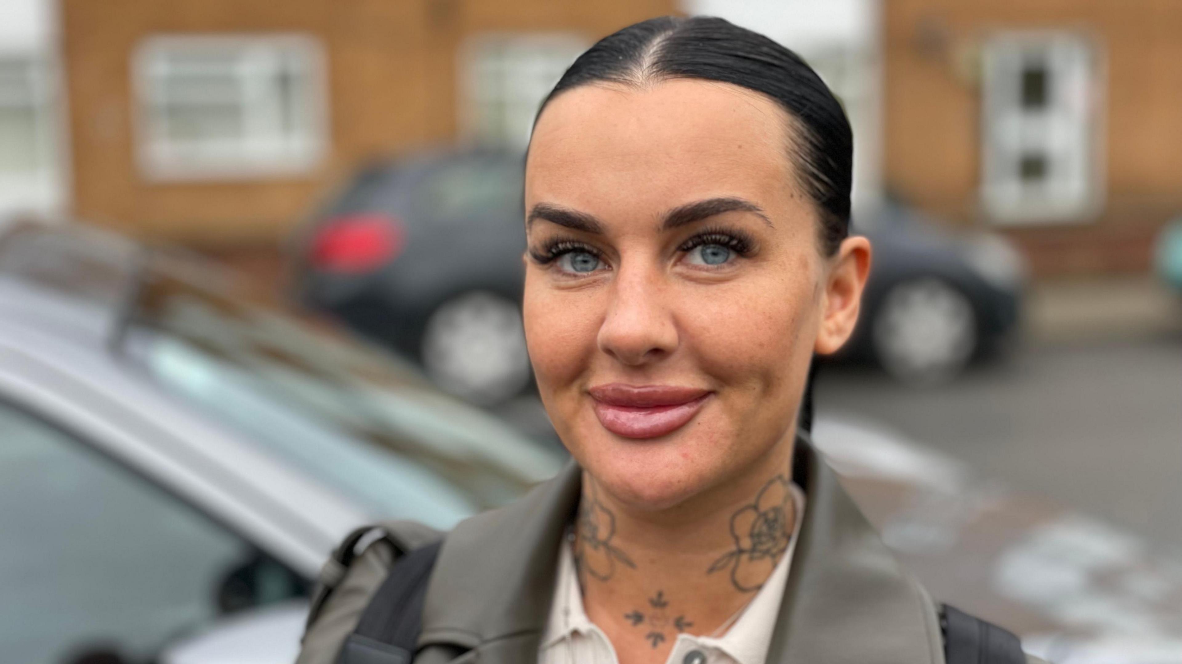 A young woman is looking into the camera lens. She has blacked hair tied back and tattoos on her neck. In the background there are cars parked in a street.
