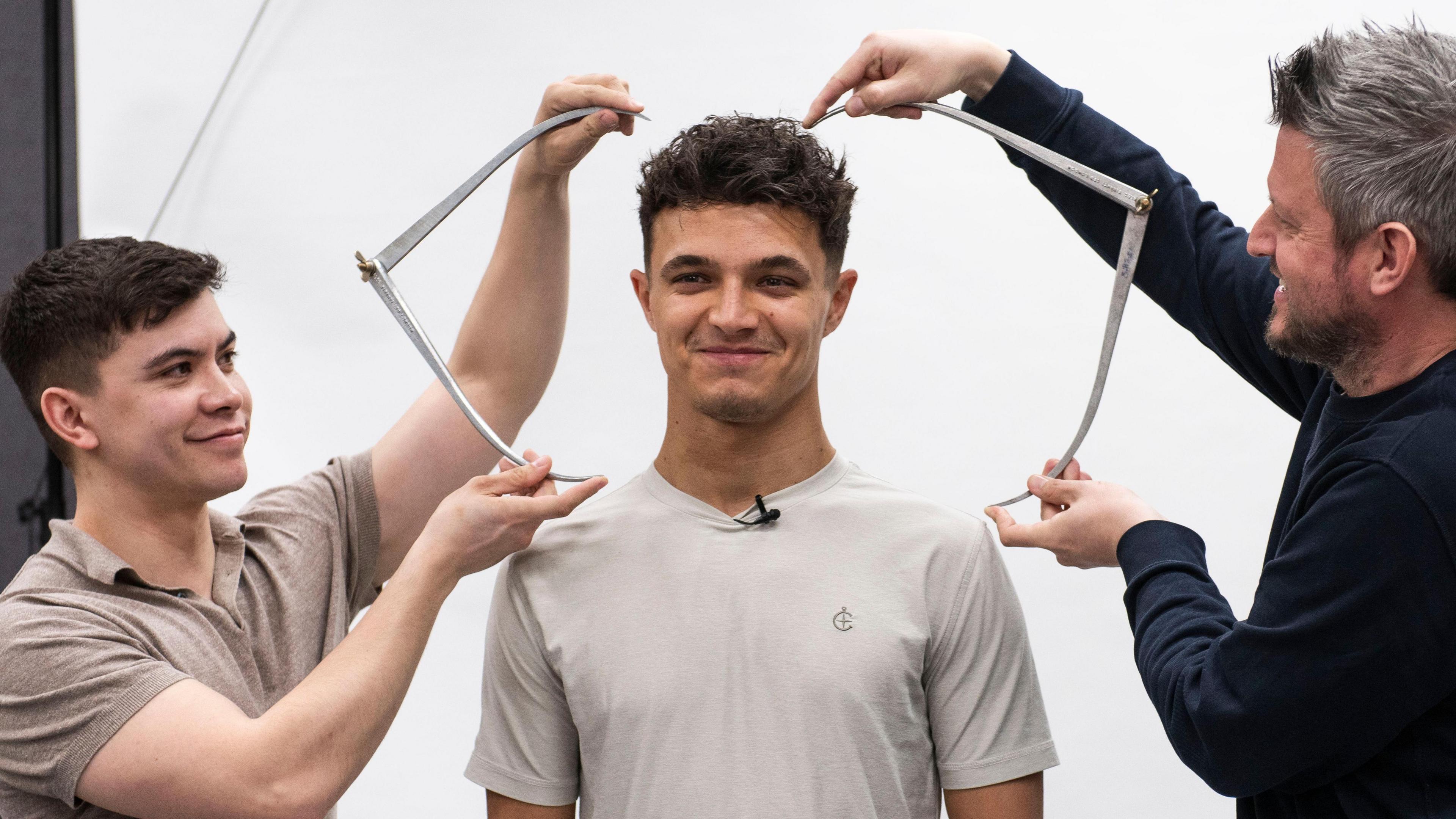F1 driver Lando Norris in a t shirt, having his head sized up by people working for Madame Tussauds.