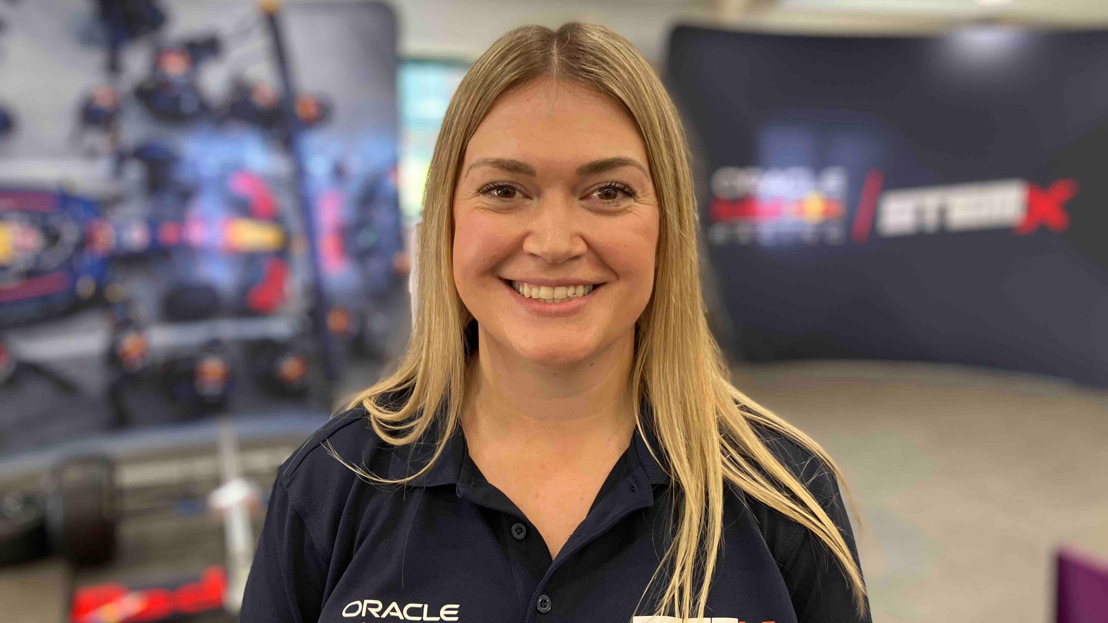 A smiling blonde woman at an event, with a disassembled F1 car behind her.