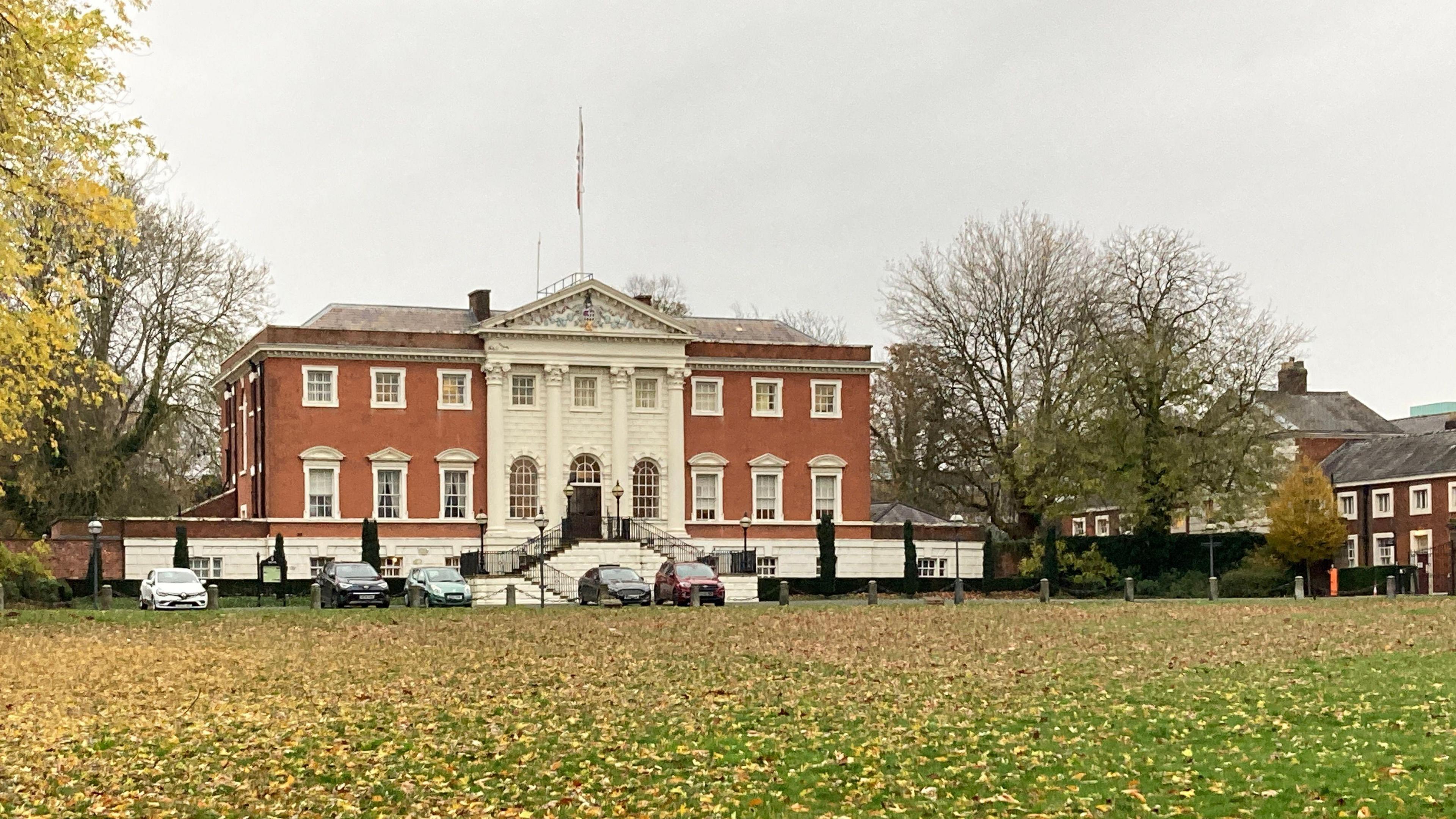 Warrington town hall stands in front of a large grassy area covered by yellow autumn leaves. The red-brick building has four white columns dominating its centre