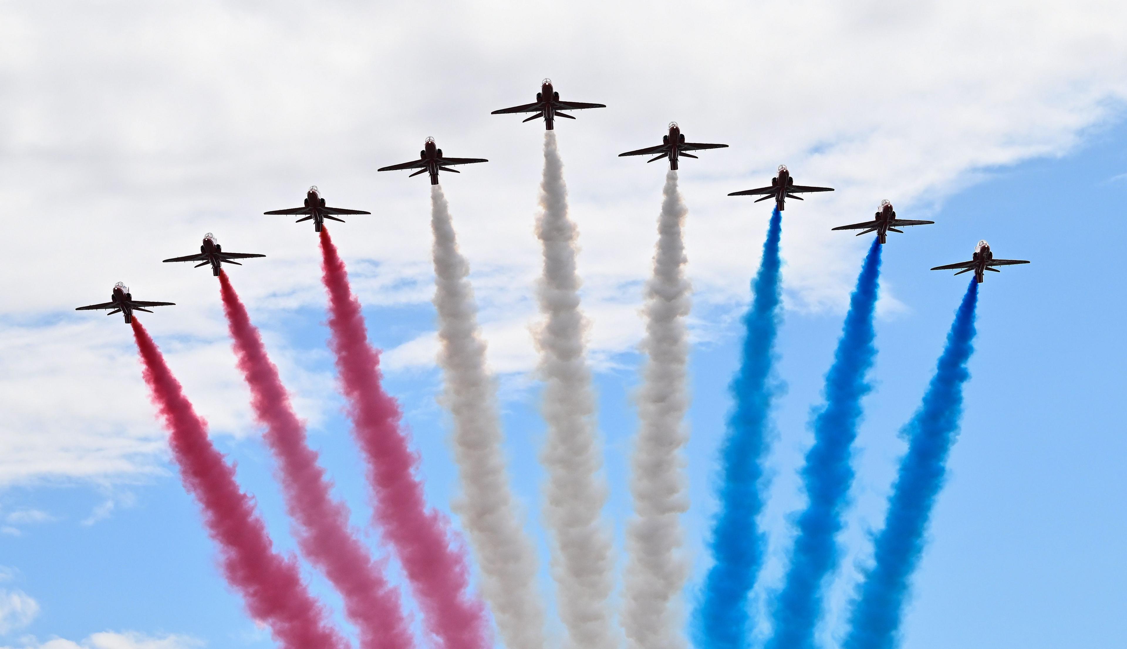 The Red Arrows flying in formation in the shape of an arrow head photographed from below. Three on the left have red smoke pouring from their tails, the middle three have white smoke and the three on the right have blue smoke. The sky is a pale cloudy blue.