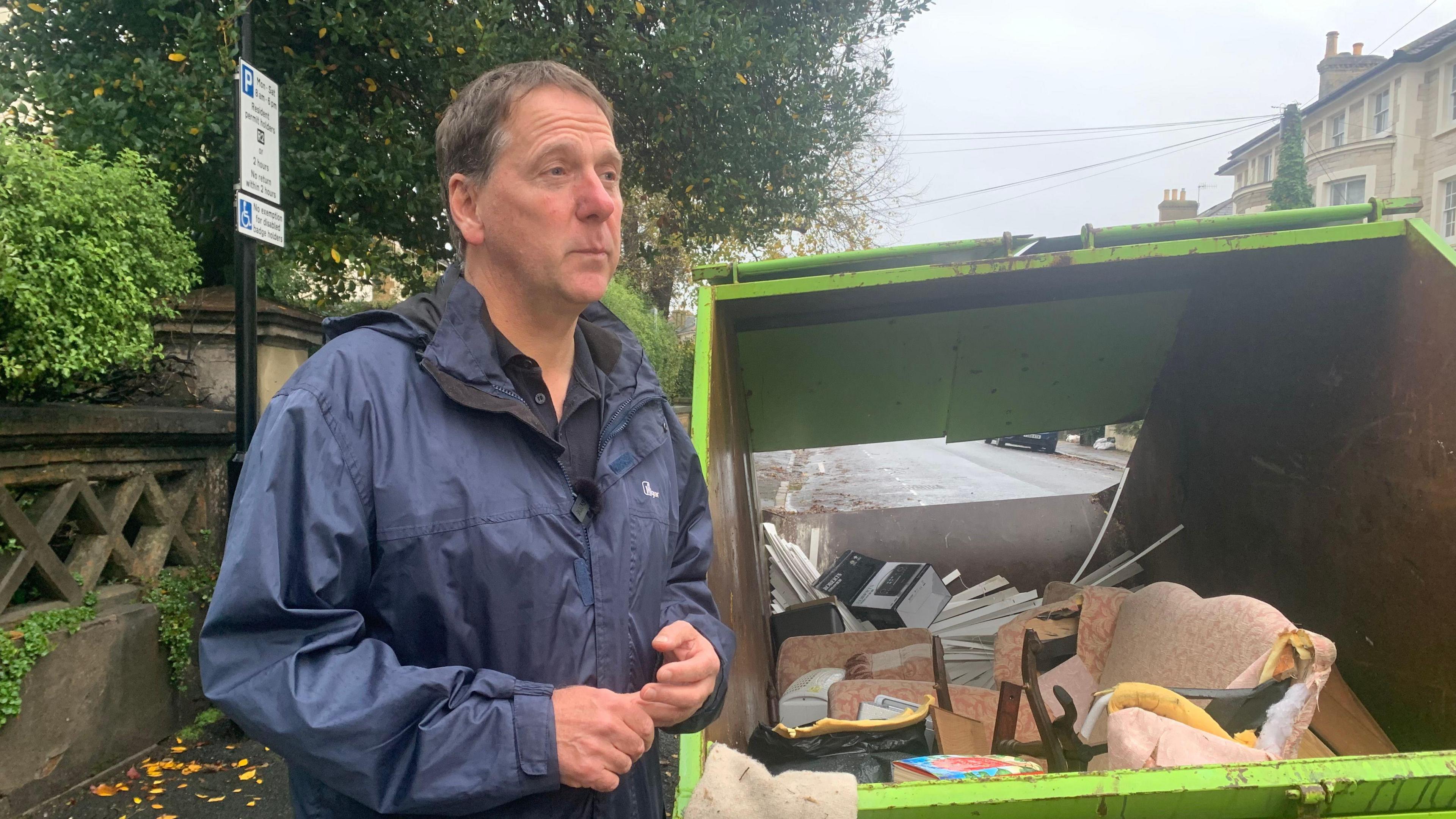 Dan Thomas, wearing a rain jacket, stands next to a skip containing ruined possessions on 8 November 2023