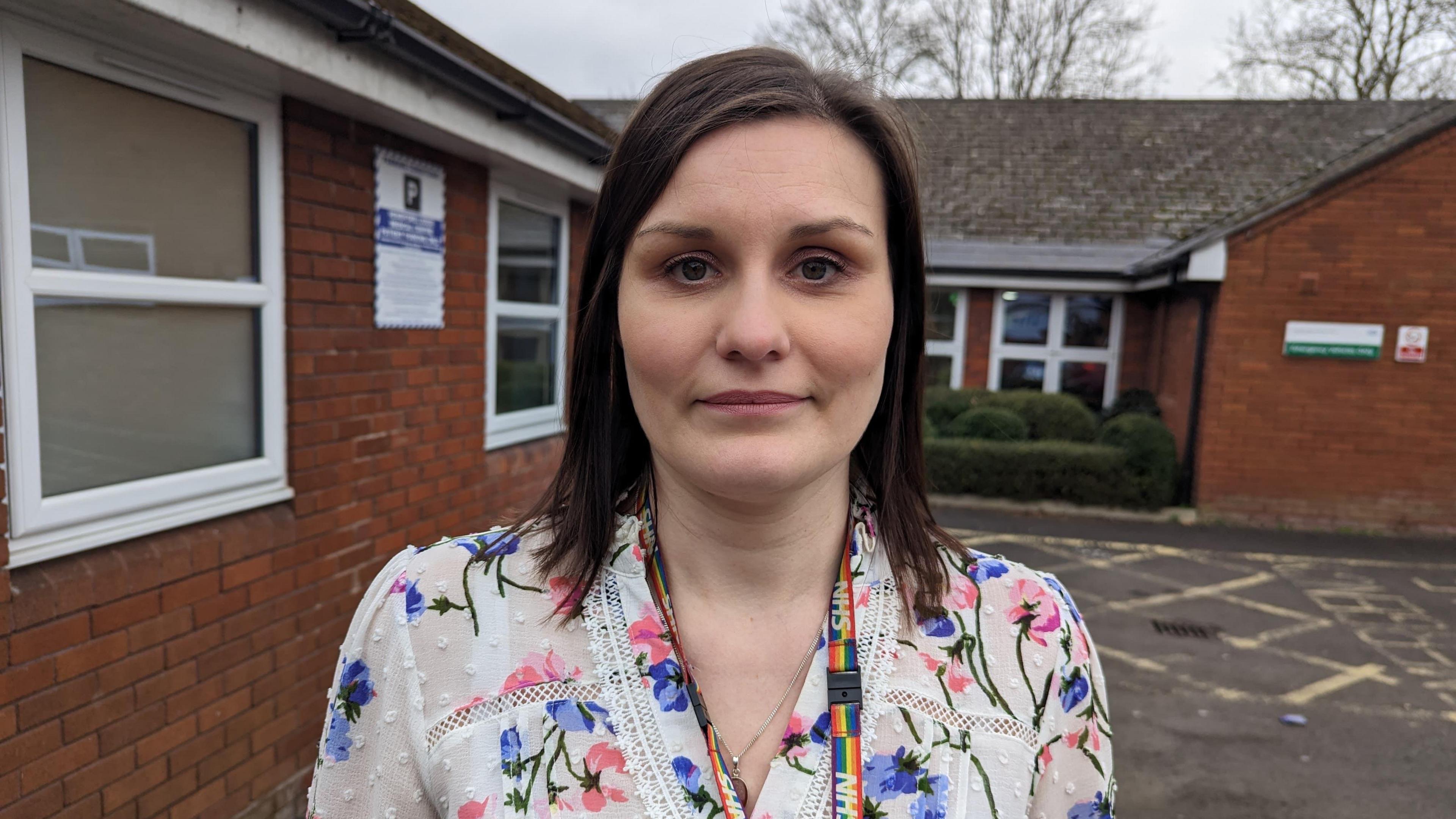 Jenna Malcolm in a white, floral top. She has dark, shoulder length hair and is wearing a rainbow-coloured NHS lanyard.