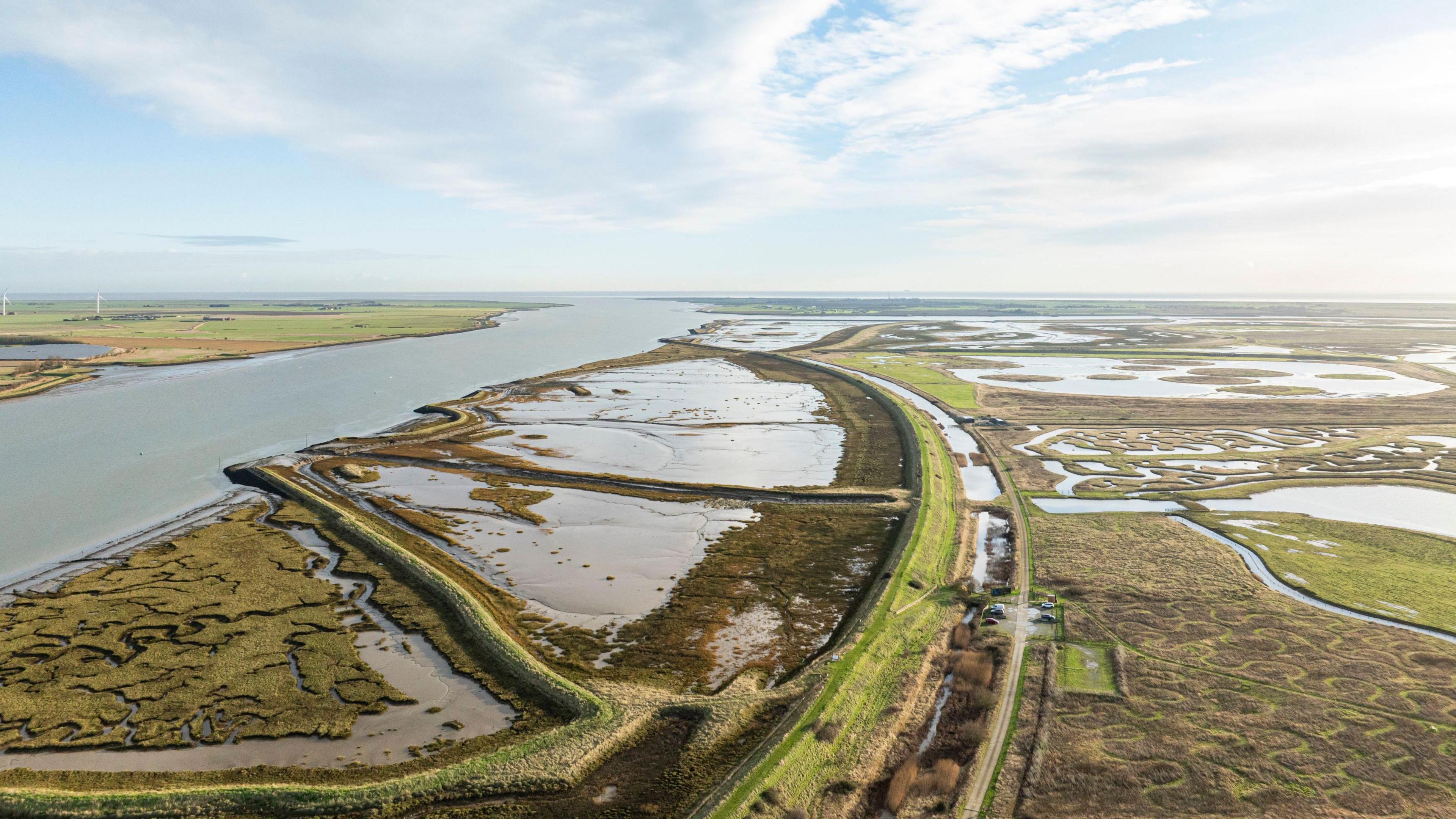 An drone view of Wallasea Island shows a thin footpath surrounded by grass, water and marsh land. The sky is blue and no buildings can be seen for miles. The area looks untouched and natural. 