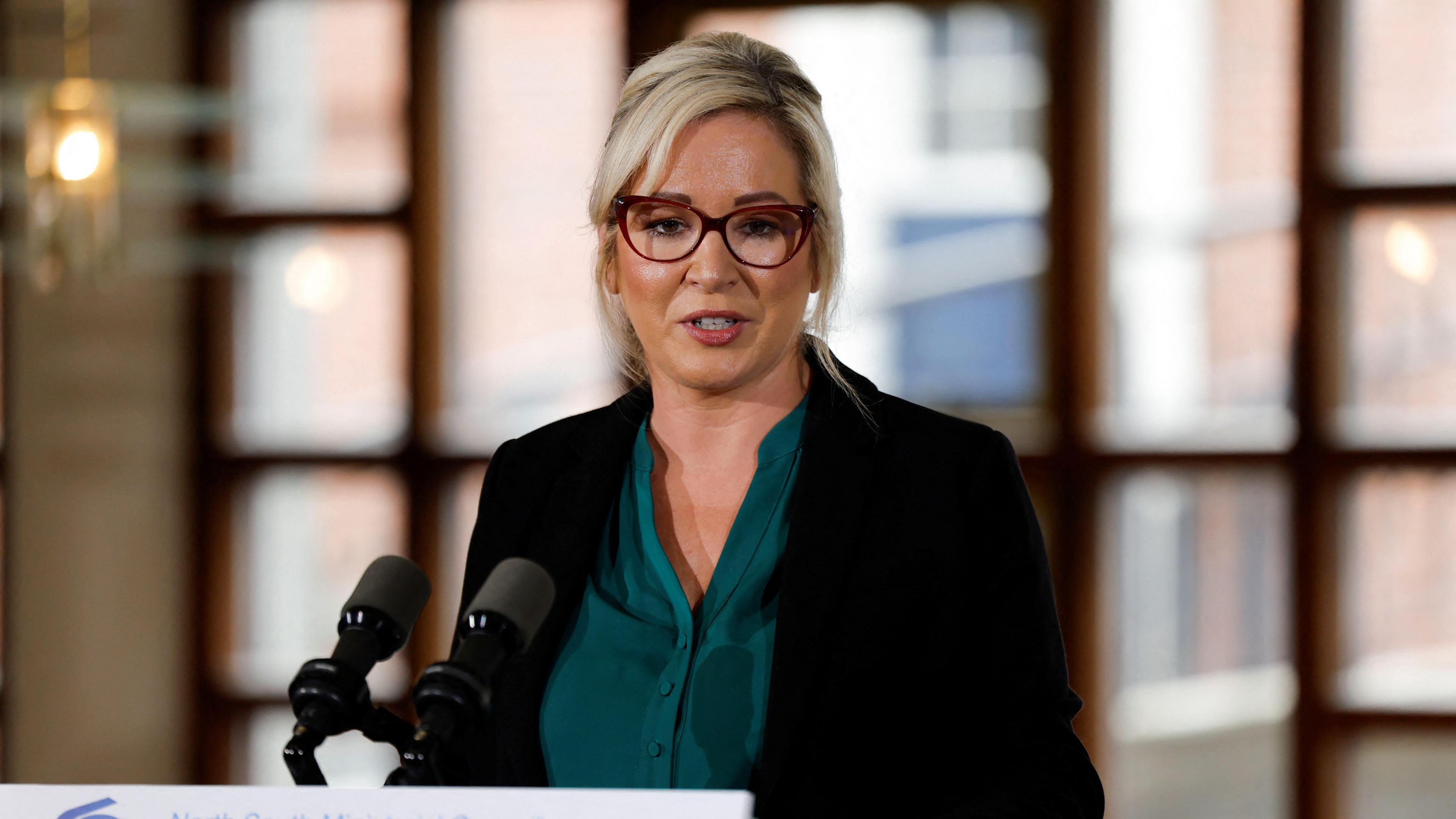 Michelle O'Neill wearing a green top and a black jacket. She is wearing glasses. 