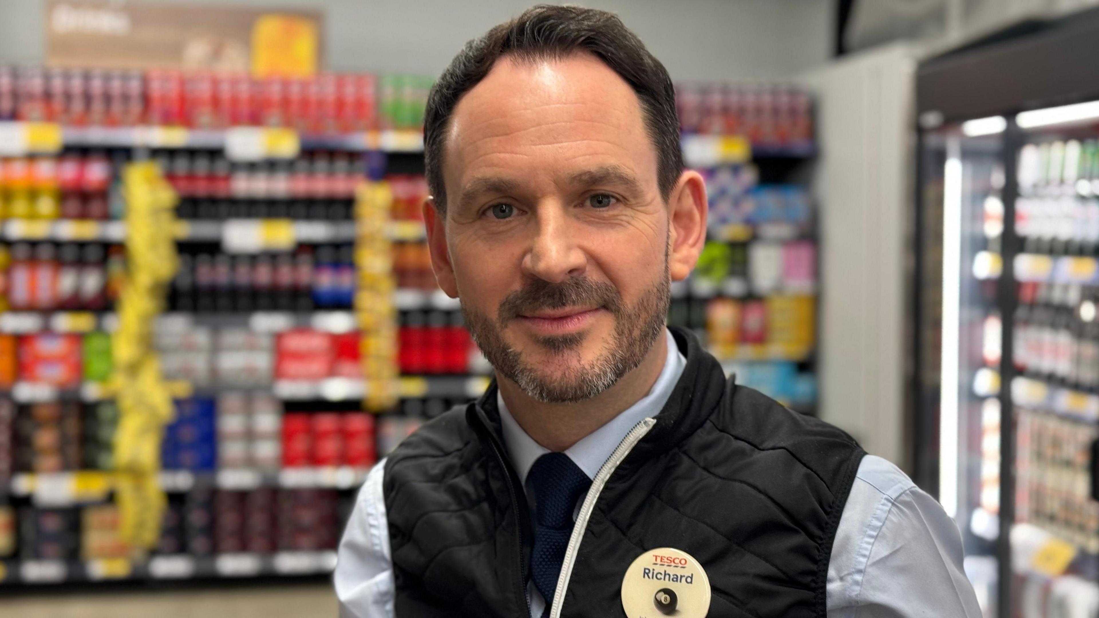 Richard Frear, who has short dark hair and a dark beard. He is wearing a navy blue gilet, powder blue shirt and name badge which says Richard. There are rows of colourful goods on the shelves behind him.