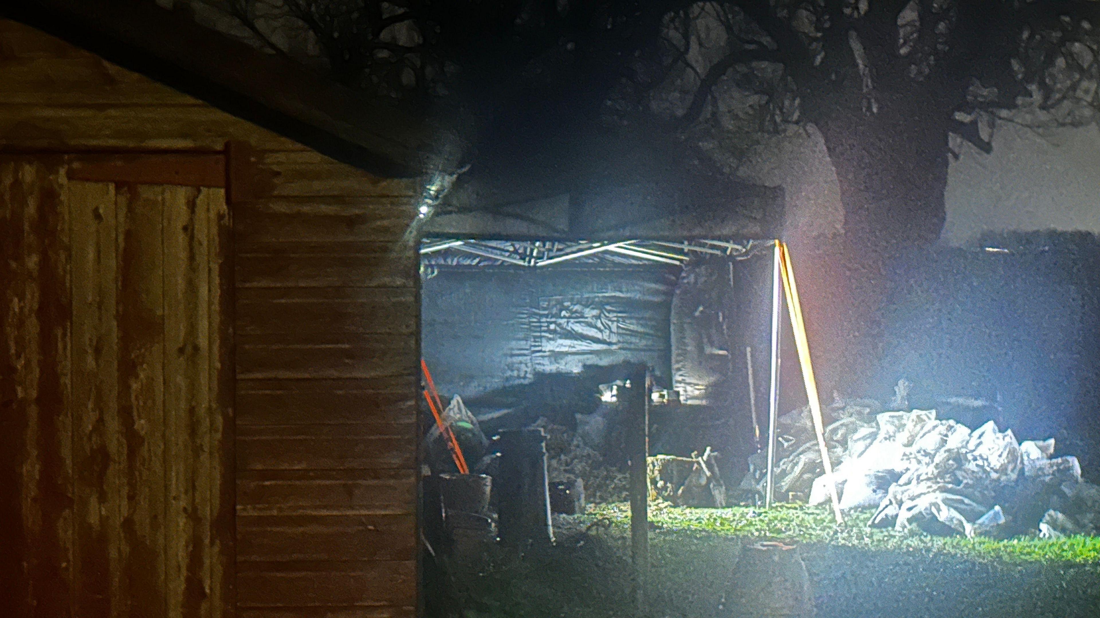 A forensic tent stands on a grassed area behind a wooden shed. A number of bags are visible on the right of the image. The sky is dark.