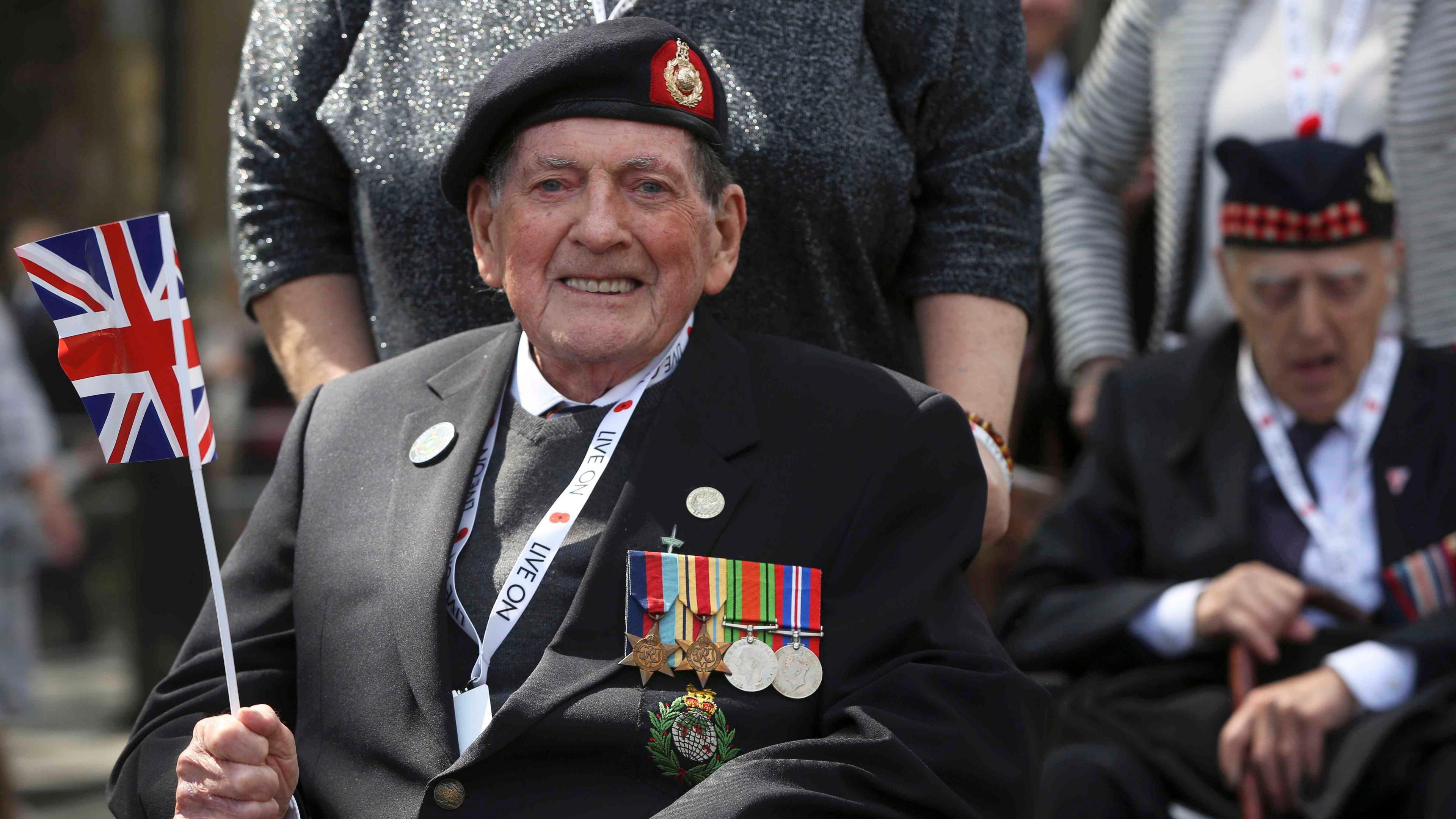 A veteran waves a Union Jack flag as he takes part in a VE Day Parade. He is wearing a suit with a line of medals across his chest.