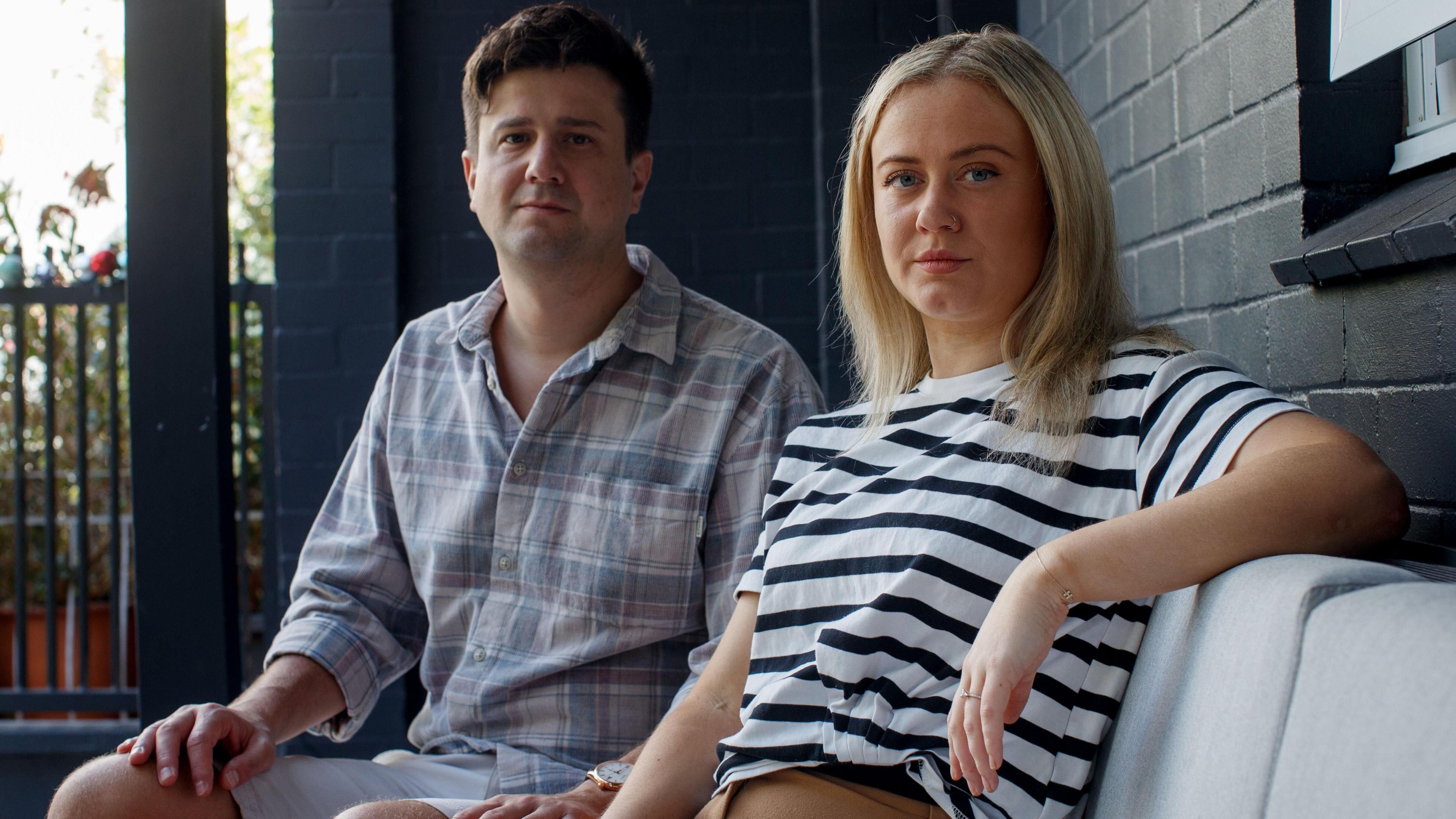 A man with short dark hair, wearing a pale blue plaid shit, and a woman with blonde hair in a black and white striped t-shirt, sit on a sofa outside a building. They are staring at the camera and are not smiling