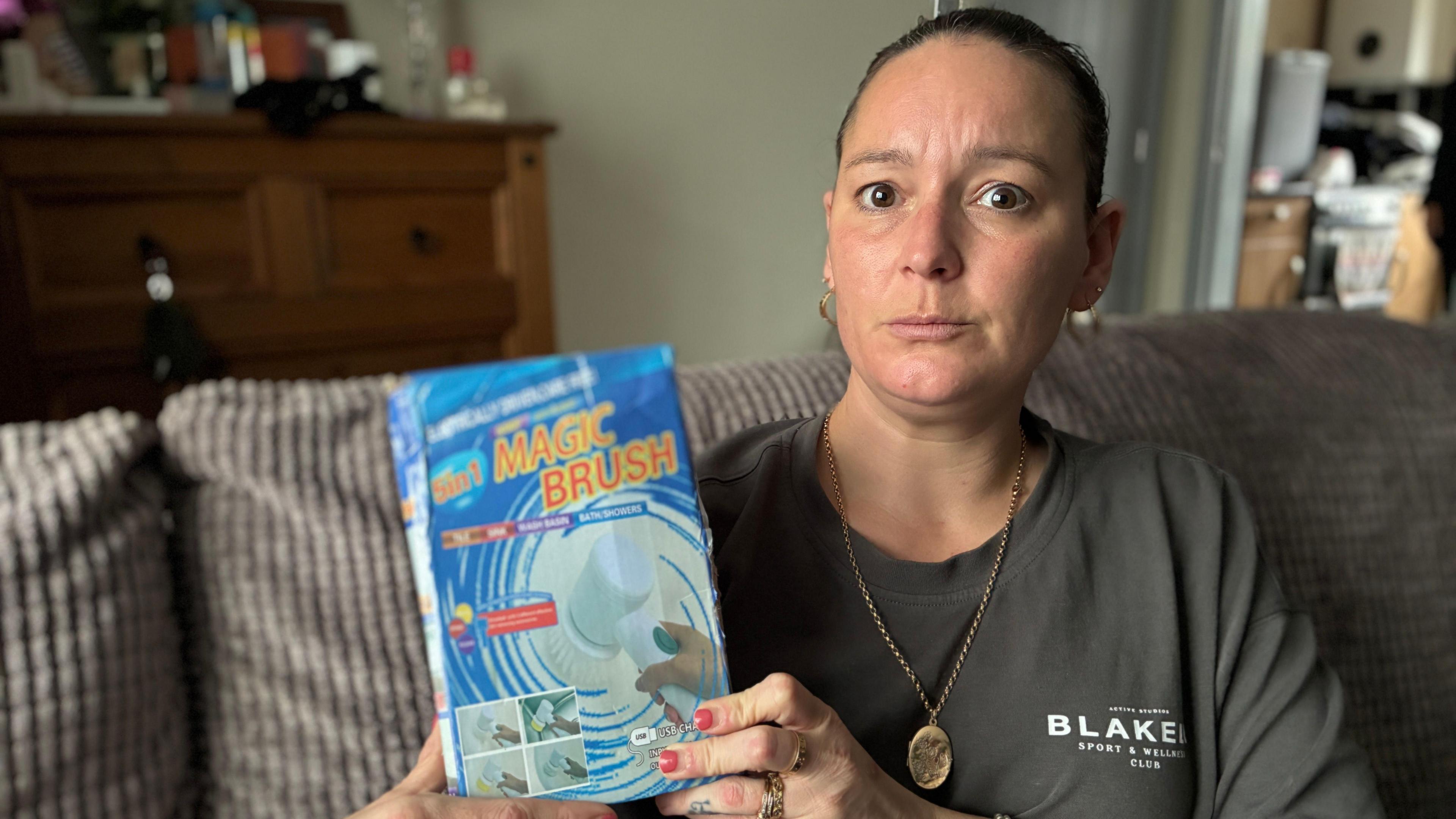 Amy is seated on a grey chair, holding the box of the cleaning brush in her hands. She has brown hair and is wearing a grey t-shirt, complemented by a gold pendant necklace.