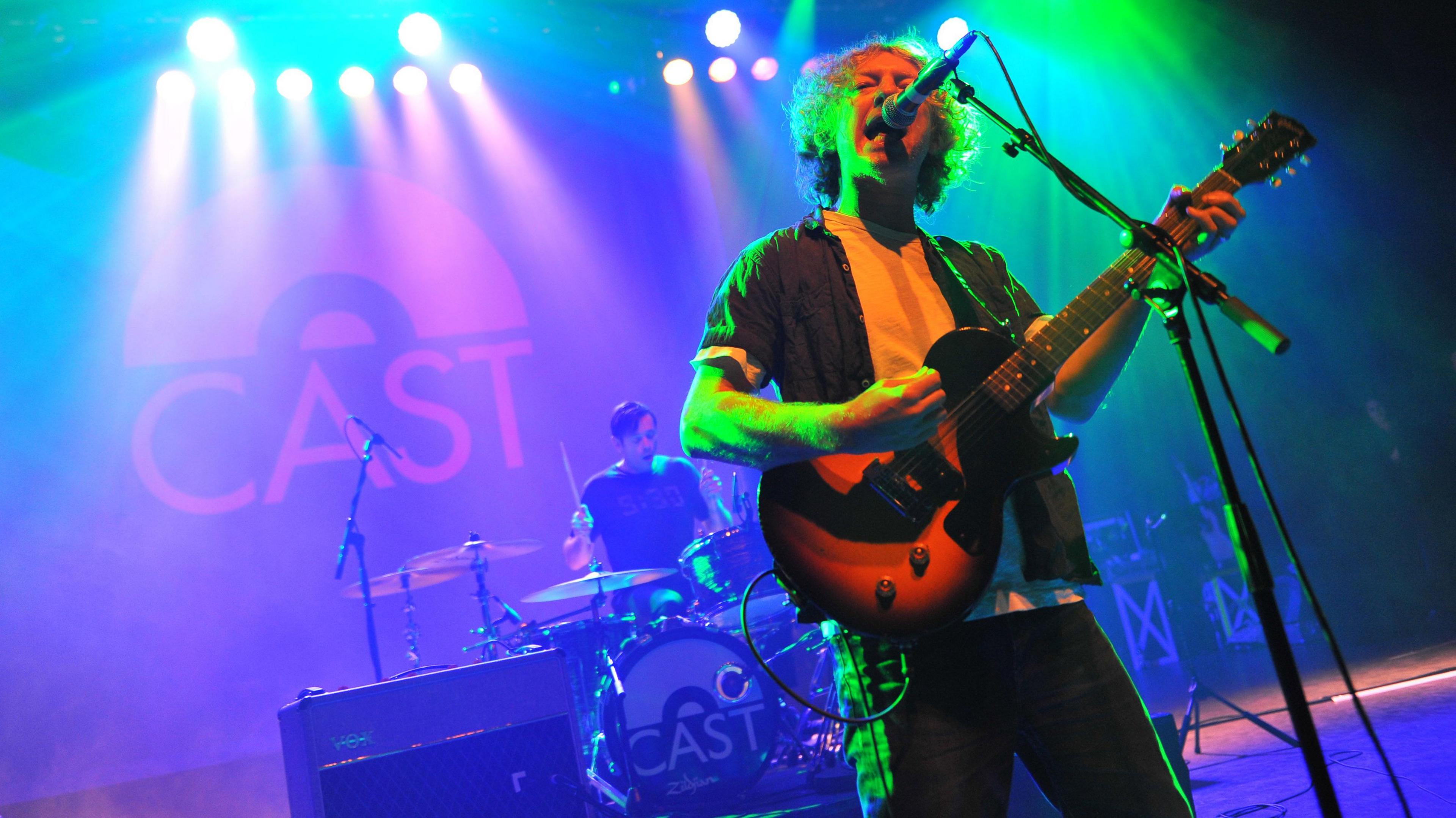 Cast's John Power sings into a microphone and plays a guitar in front of a drummer and a large backdrop with the word "Cast" on it