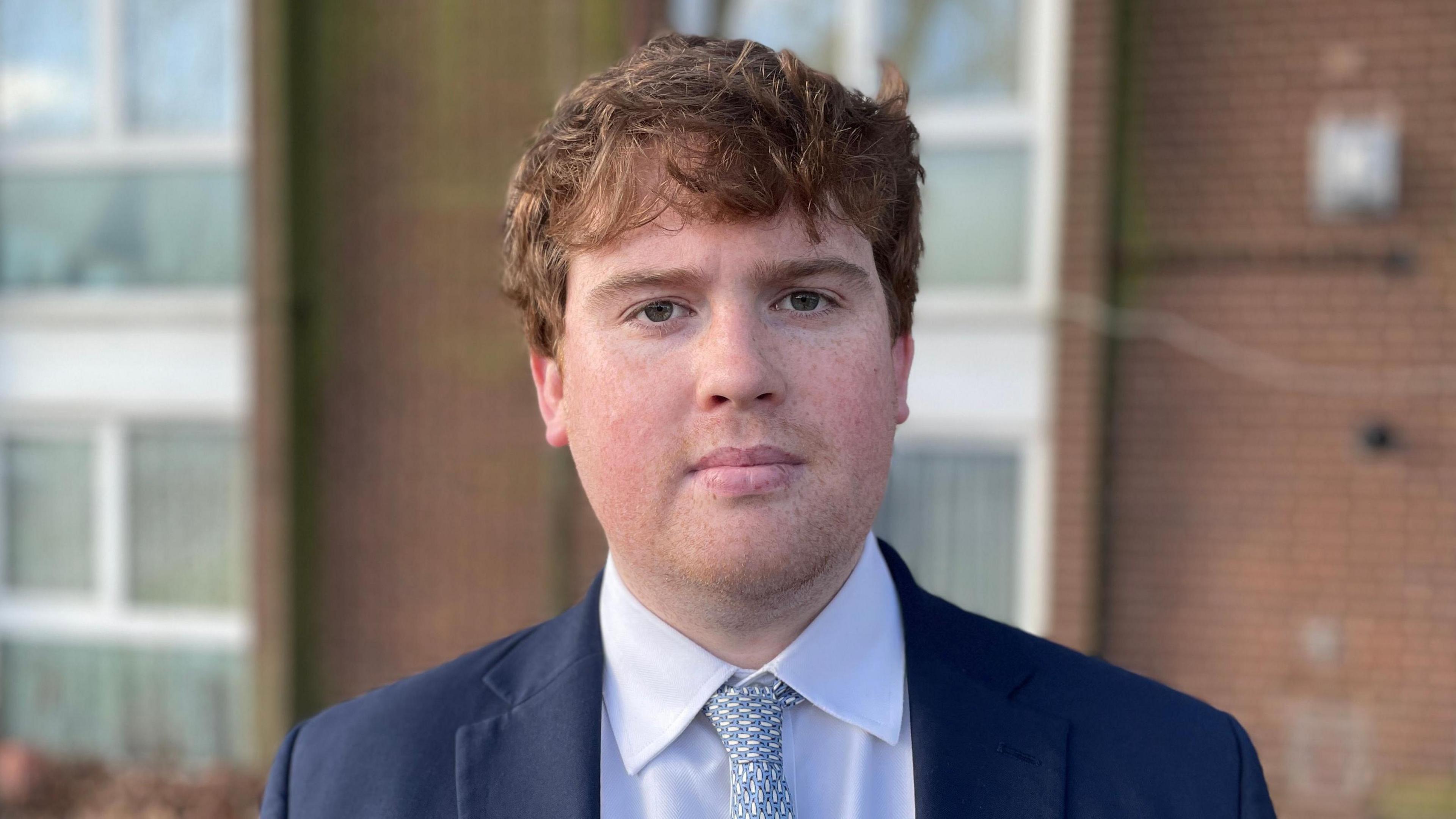 Dan Swords, who has medium length, light brown hair. He is standing in front of a block of flats with a serious expression on his face. He is wearing a navy suit jacket, white shirt and light blue tie.