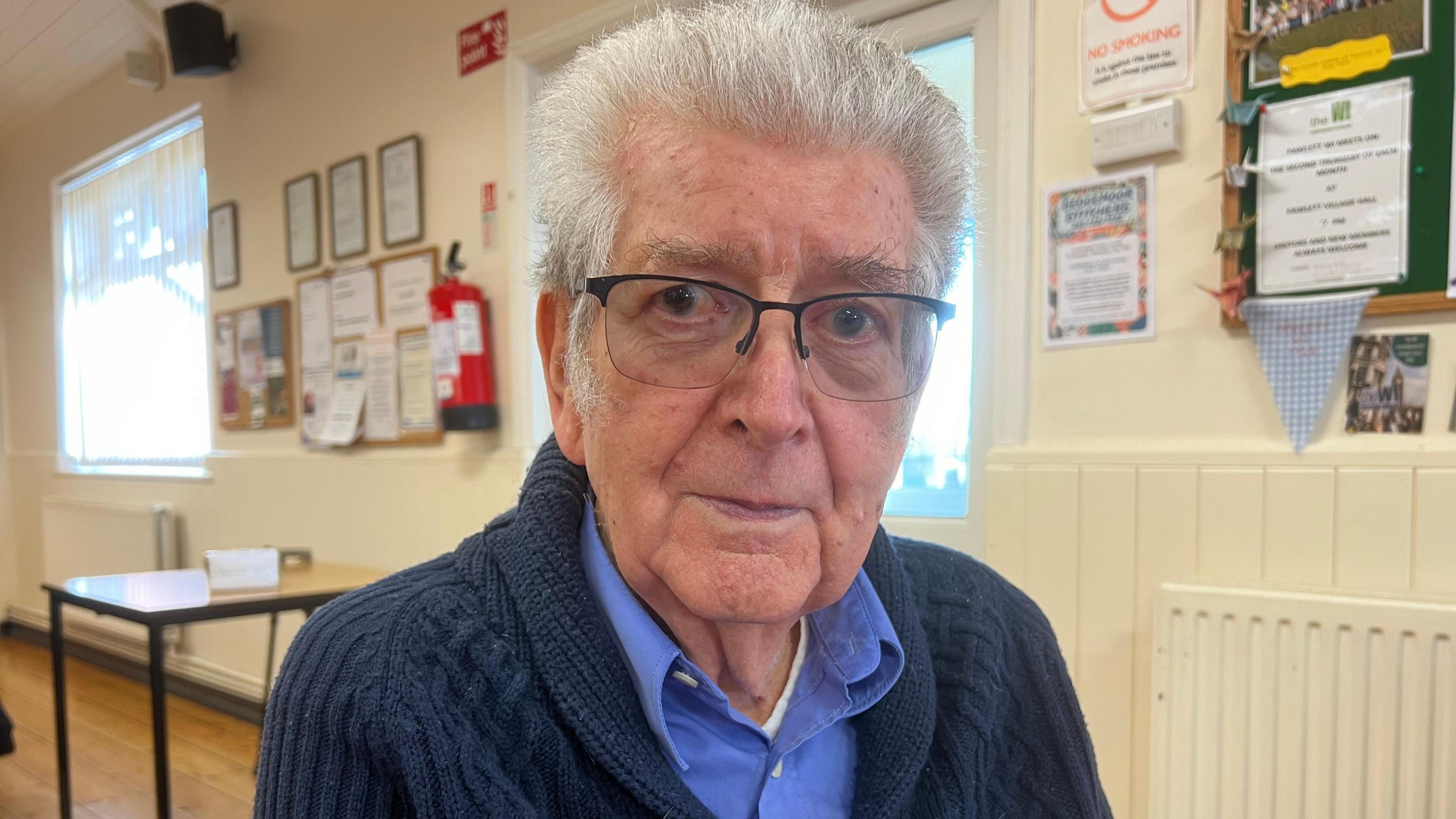 Tony Wadsworth. He has short white hair and glasses. He is wearing a blue shirt under a navy blue knitted cardigan. He is looking at the camera and smiling. 