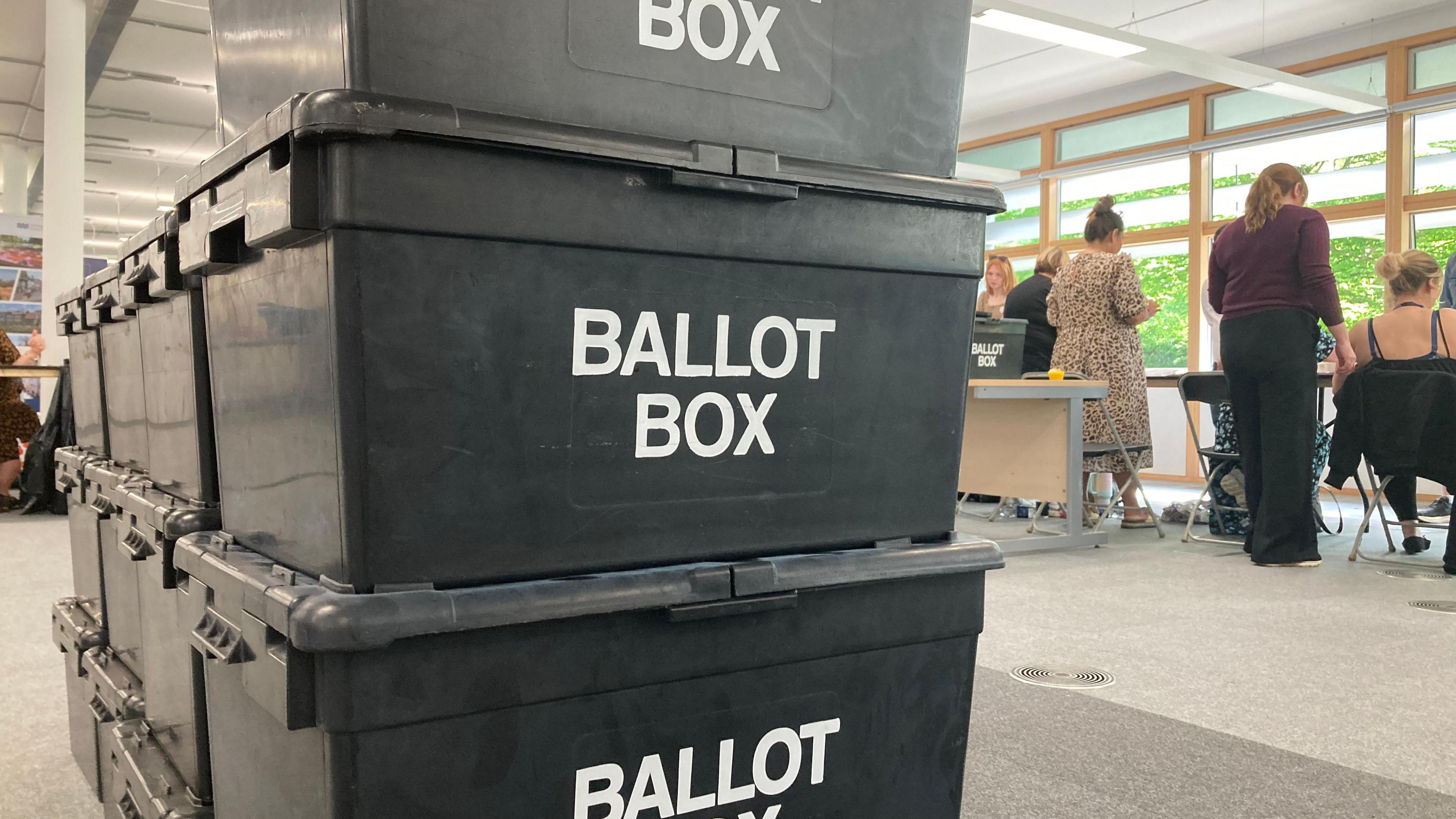 Three black boxes stacked on top of each other with the words 'Ballot Box' printed on the front in white. In the background people are counting ballots.