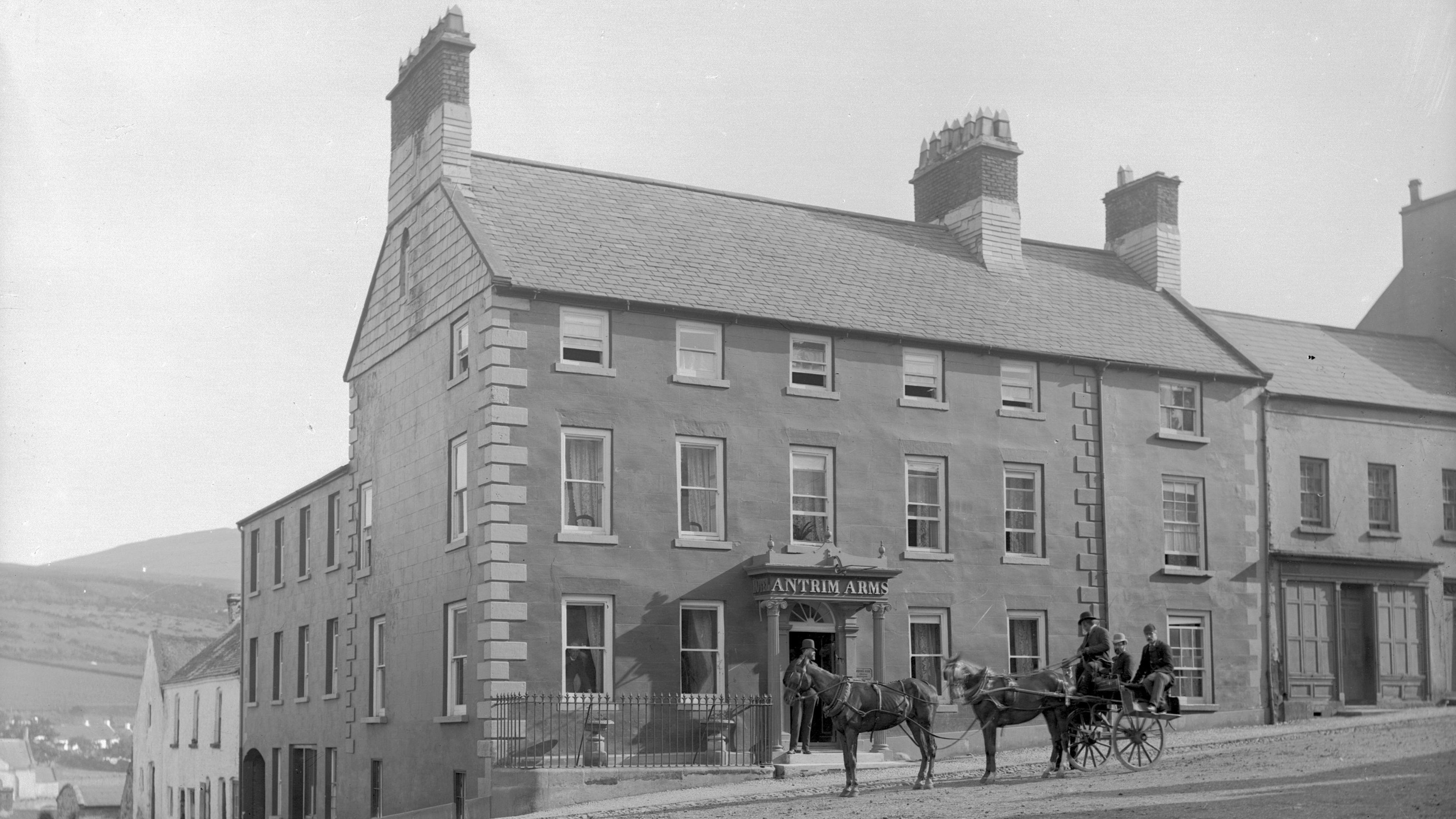 A black and white archive photo of the Antrim Arms hotel in Ballycastle.   The hotel is a large Georgian-era building on a corner site.  There is a cart pulled by two horses waiting outside the entrance.  Three well-dressed men are sitting on the cart. 