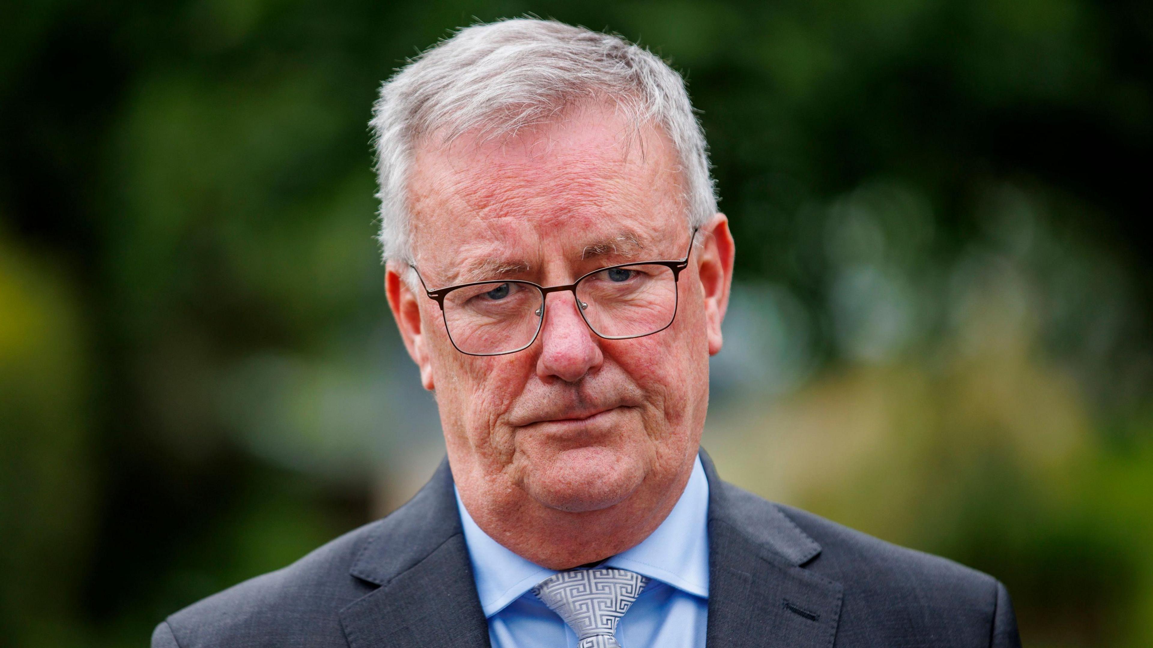 Mike Nesbitt. He has short grey hair, wearing glasses, a dark grey blazer, blue shirt and silver tie.